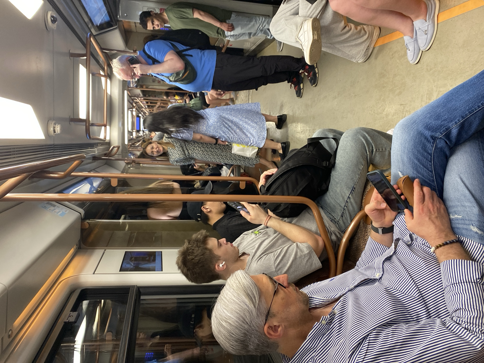 The image shows the interior of a passenger train, likely a subway car. The scene includes several people, mostly passengers, who are in various states of activity. Some are standing, while others are seated. Many of the passengers are looking at their phones. The train is illuminated with artificial light. Based on visual cues, there is no indication this image is necessarily from Moscow, Russia.
