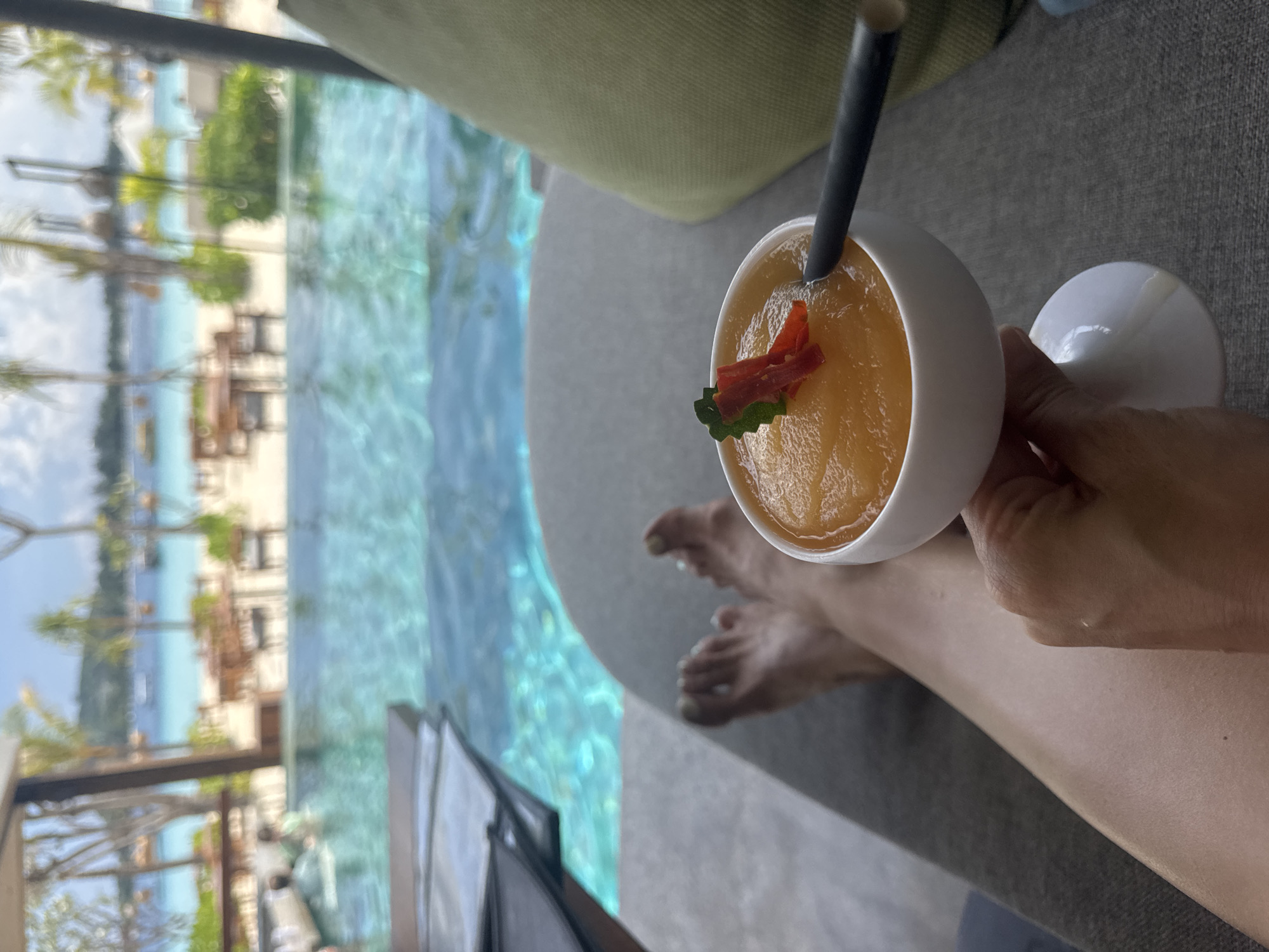 A person's legs and feet are seen next to a white stemmed glass holding an orange smoothie garnished with a sprig of mint and a red fruit or vegetable slice. A black straw is inserted in the smoothie. The glass is held by a hand.
Outside, there is a view of a partially visible turquoise pool and a blurred beach scene with seating, foliage, and buildings under a bright sky. The location is presumed to be in a resort, possibly in Unknown, Indonesia.