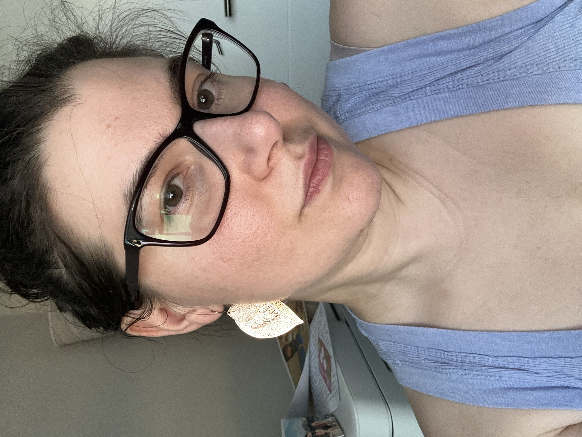 The image is a close-up, portrait-style photograph of a woman wearing glasses and a light blue tank top. She has dark hair pulled back loosely and is wearing large, decorative leaf-shaped earrings. The lighting suggests an indoor setting, possibly near a window. The background is slightly blurred, showing what appears to be a desk or counter with some papers and objects on it, and a pale blue wall. There are no discernible landmarks or contextual clues to indicate a specific location in Leeds, United Kingdom. The focus is on the woman's face and upper body.