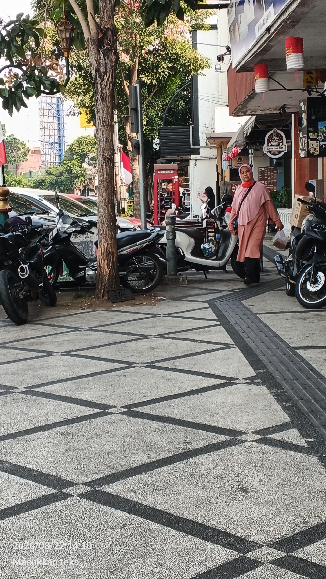 The image depicts a street scene in Malang, Indonesia, likely on a sidewalk adjacent to a building. In the foreground, a woman wearing a peach-colored hijab and dress is walking towards the right side of the frame, carrying a plastic bag. She appears to be a pedestrian on the sidewalk. Behind her, several motorcycles are parked along the roadside, suggesting this is a common area for parking. To the left, there are cars parked as well. The environment includes trees with foliage, and part of a building with signage and some decorative elements, possibly indicating a commercial area. The sidewalk itself has a textured pattern of dark lines on a light gray surface. The presence of a red telephone booth and Indonesian flags in the background further confirms the location in Indonesia. The overall scene is of a typical urban street with pedestrians, vehicles, and commercial establishments.