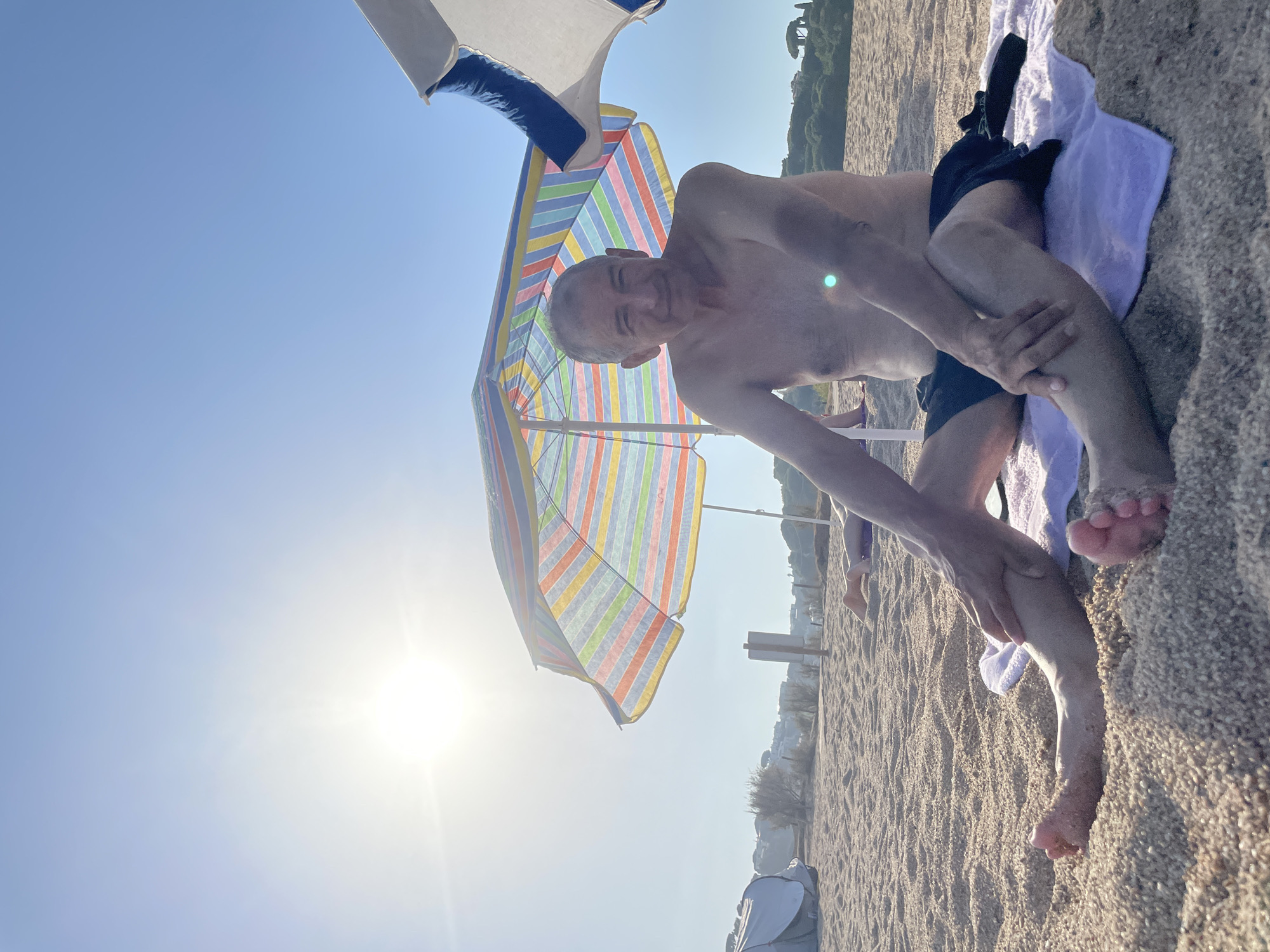 An older man with graying hair and a bare torso is sitting on a sandy beach in Canet de Mar, Spain. He is wearing dark swim shorts and is seated on a light purple towel. His legs are crossed, and his left hand rests on his right shin. To his left is a colorful striped beach umbrella, and to his right, a white umbrella is partially visible. The sun is shining brightly in the clear blue sky, casting a lens flare on the man's chest. The beach sand is granular, and other beachgoers are visible in the background, along with some vegetation and buildings on a distant hill. A pop-up tent is also present on the left side of the image.