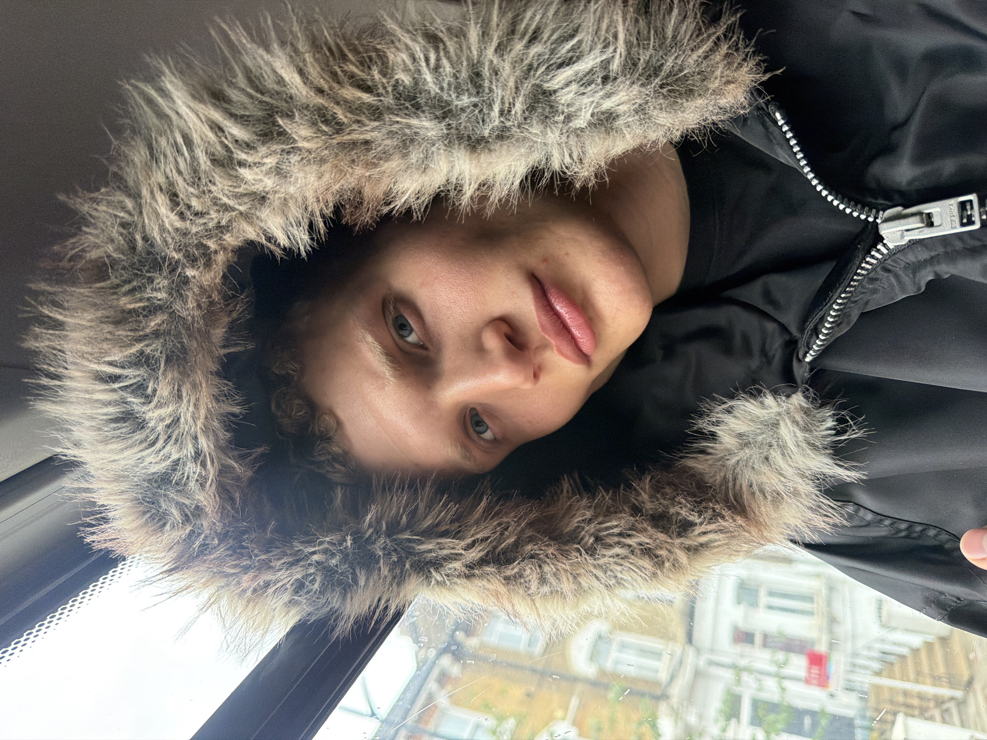 A young person, with fair skin and blonde curly hair, is seen in a close-up selfie. They are wearing a black hooded jacket with a prominent faux fur trim around the hood. The person's head is tilted slightly, and they are looking directly at the camera. Their expression is neutral. The background suggests they are inside a vehicle, possibly a bus or train, as evidenced by the window and the reflection of buildings on the exterior. The buildings appear to be terraced houses, typical of a residential street in London. The window shows a slightly overcast day, with rain streaks visible on the glass.