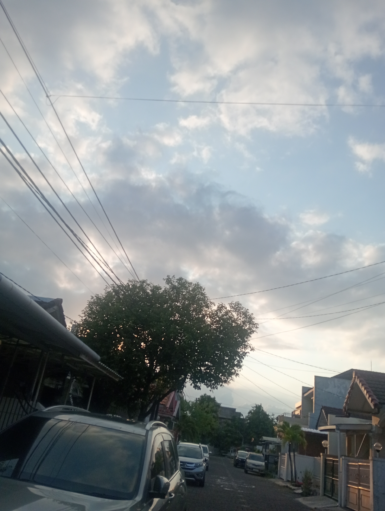 The image depicts a street scene in Surabaya, Indonesia, at dusk. The sky is filled with a mix of clouds and patches of blue, illuminated by the setting sun, creating a soft, diffused light. Overhead, multiple electrical wires are strung across the frame, a common sight in urban Indonesian settings.
In the foreground, the front of a silver SUV is visible, its roof rack prominent. To the left, a portion of a building with a corrugated metal roof is seen. Further down the street, several other vehicles are parked and driving, including a blue hatchback and other cars. Residential buildings with modern architectural styles line the right side of the street, featuring tiled roofs and gates. Lush green trees are scattered throughout the scene, with one particularly large tree with dense foliage positioned centrally behind the SUV. The road surface is asphalt. The overall impression is one of a quiet residential street as day transitions into evening.