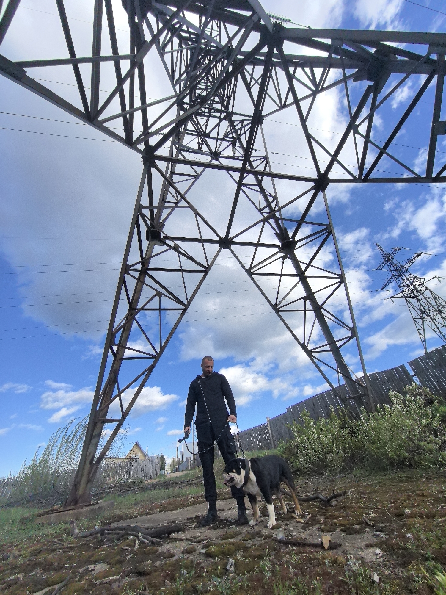 A low-angle, full-shot image depicts a man and a dog standing beneath a large electrical transmission tower. The scene is located in the city of Noviy, Russia.
The man is wearing a black jumpsuit and black boots. He is holding a black leash attached to a black and white English Bulldog. The dog stands with its head slightly lowered.
Dominating the background is a massive, metal electrical transmission tower, with another similar tower visible further in the distance to the right. Overhead, high-voltage power lines run from tower to tower, against a backdrop of a blue sky dotted with fluffy white clouds.
The ground is overgrown with vegetation, including moss, grass, and small bushes. In the middle distance, a weathered wooden fence runs alongside the power lines. Some small, wooden buildings can be seen over the fence. The light suggests it is daytime.