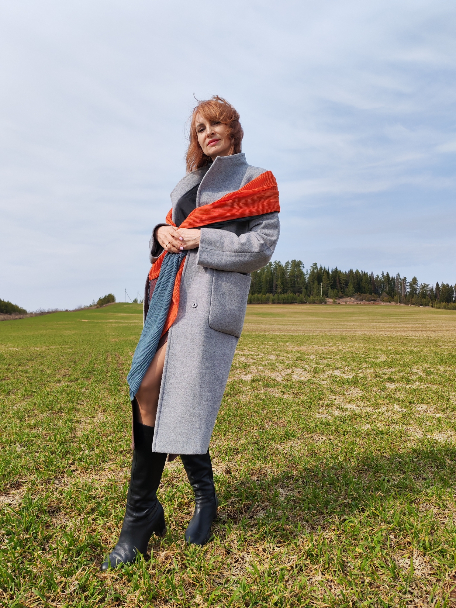The image depicts a woman standing in a field, likely located near Нечкино, Russia based on the presence of the Нечкинский национальный парк (Nechkinsky National Park). The woman has reddish-brown hair and is wearing a grey coat, black boots, and a pleated scarf with alternating orange and blue colors. She is positioned diagonally in the frame, facing the camera, with her hands clasped in front of her.
The field is covered with short green grass mixed with some dry patches of ground. In the background, there is a line of trees, suggesting a forest or wooded area. The sky is a light blue with scattered white clouds.