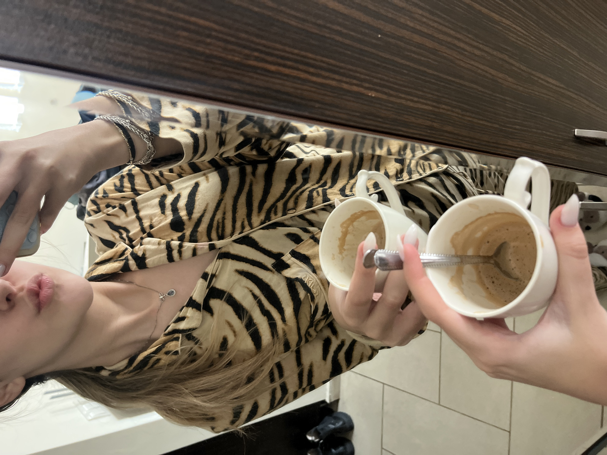 The image is a selfie of a young woman with long blonde hair, light skin, and pink lips, wearing a tiger-striped shirt, taken in a mirror. She is holding two white ceramic coffee cups, one in each hand. The cup in her right hand contains a spoon in it. She is wearing a silver necklace and multiple silver bracelets on her left wrist. Behind her, there's a dark brown wooden cabinet and a tile floor. Based on the generic description, it is impossible to determine if the location is in Sofia, Bulgaria. More information would be needed.