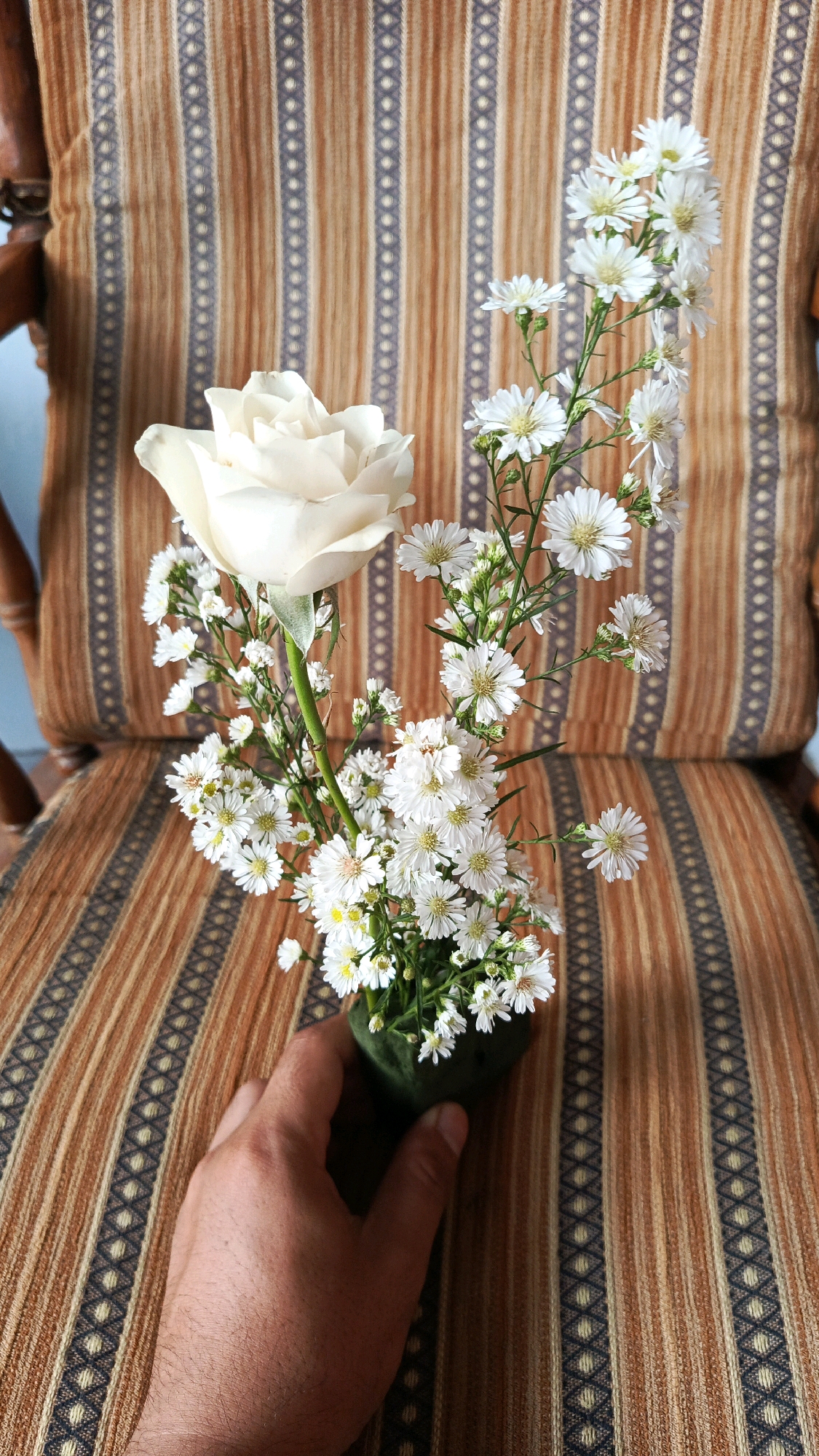 A close-up shot shows a hand holding a small bouquet of white flowers, including a prominent white rose and smaller white aster-like flowers, in a dark green holder. The bouquet is positioned on a striped fabric background, which appears to be upholstery on a chair. The fabric features alternating vertical stripes of brown and tan, with a repeating pattern of small, diamond-like shapes in a dark blue or black color within some of the tan stripes. The scene is lit from above, creating some shadows. The background also shows a hint of dark wood, likely part of the chair's frame, and a pale blue wall. The location is identified as Babadan, Indonesia.