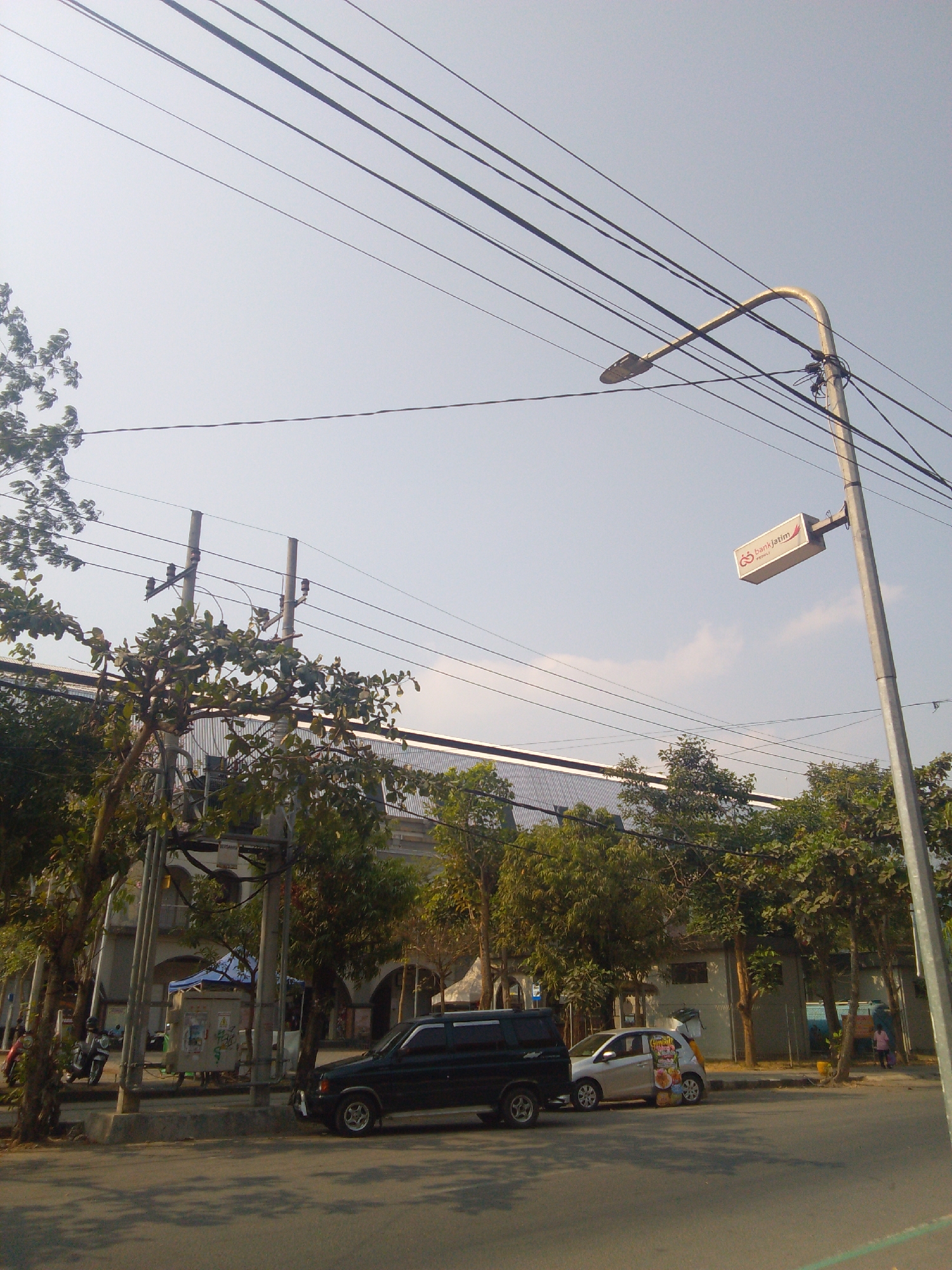 The image depicts a street scene in Trenggalek, Indonesia. The foreground is dominated by a paved road with dappled sunlight and shadows. A dark colored SUV is parked on the right side of the road, facing towards the left. Next to it, a lighter colored car is also parked. In the background, several trees with green foliage line the street. Behind the trees, a long, modern-looking building with a metallic roof is visible. Overhead, numerous power lines crisscross the sky. A street lamp with a curved arm extends into the frame from the right, and a rectangular sign for "bankjatim" is attached to it. Further to the left, there are utility poles supporting more power lines, and part of a multi-story building with arched entrances can be seen. In the middle ground, near the utility poles, there are what appear to be small stalls or kiosks, one with a blue tarp. A red motorcycle is parked to the left of these stalls. A few people are visible in the scene, including one person walking on the sidewalk in the distance on the right, and another person near the stalls on the left. The sky is clear with a light haze.