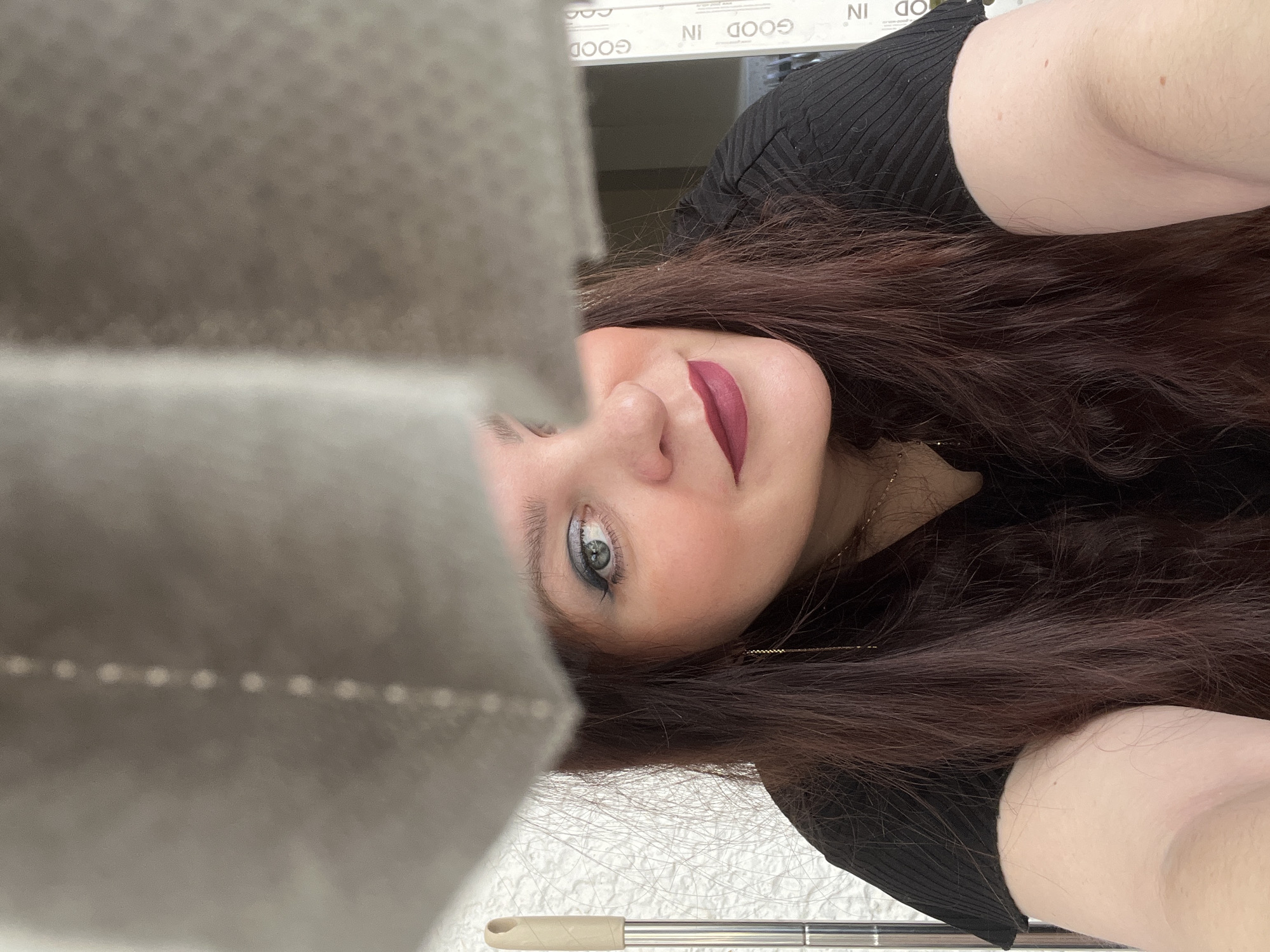A selfie photograph features a fair-skinned woman with auburn hair wearing a black, short-sleeved top and a dark lip color. She has blue-gray eyes with dark eyeliner. A gold necklace is visible on her neck. The background behind her includes the words "GOOD IN" repeated on a white frame. A fuzzy gray object with small white dots is in the foreground. The bottom of the image includes a mop and a white textured surface. The photo does not provide enough information to determine the location in Noviy, Russia.