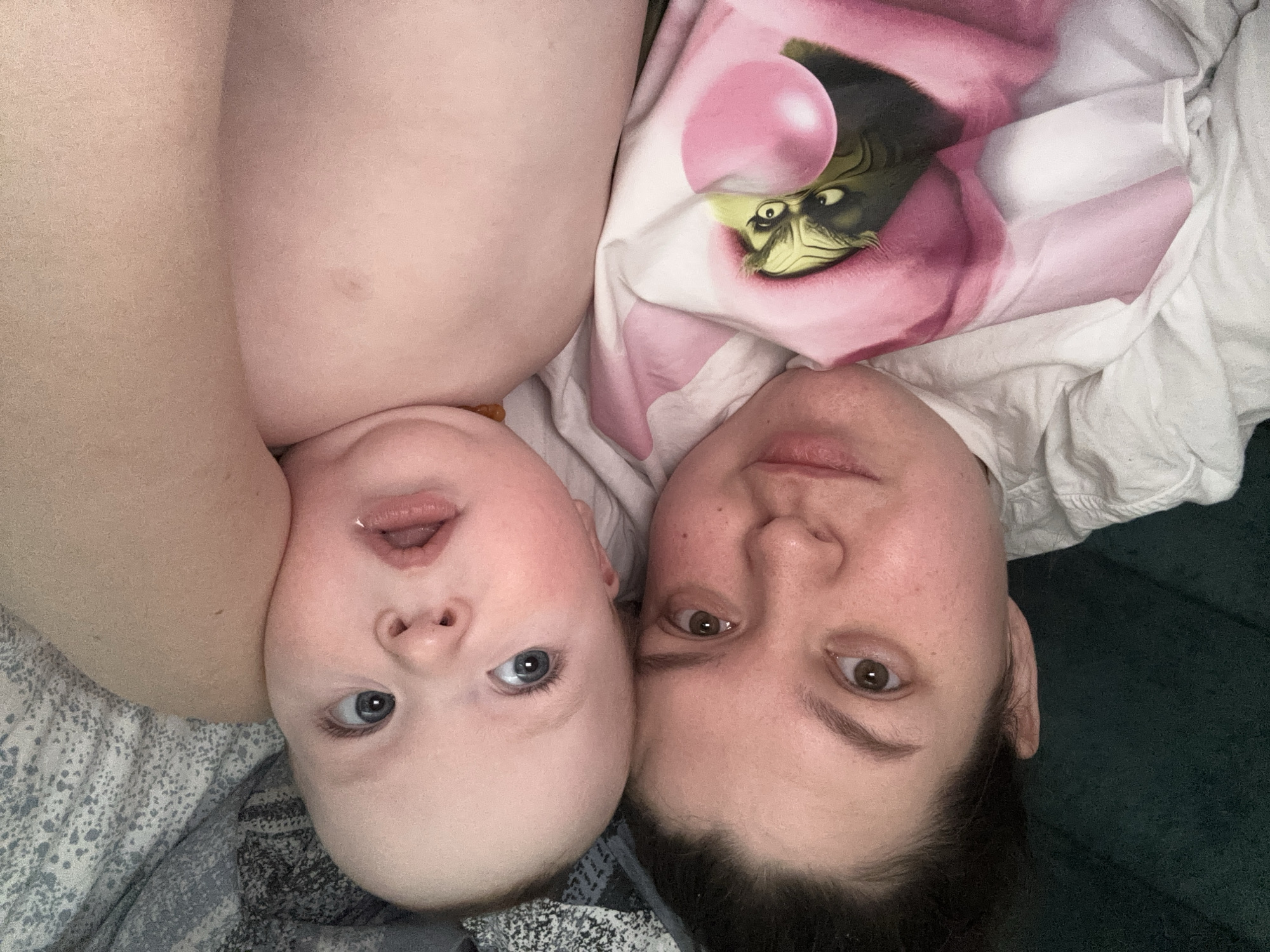 A close-up, downward-angle shot features a baby and an adult lying down next to each other. The baby, on the left, has fair skin, blue eyes, and an open mouth. The adult, on the right, has fair skin, brown eyes, and dark hair. Both are facing upwards towards the camera. The adult is wearing a white shirt with a pink graphic featuring the Grinch. The baby is lying on a patterned grey and white blanket, while the adult is lying on a dark green surface. The location is likely indoors. The city, Марковский, Russia, is not identifiable within the image.
