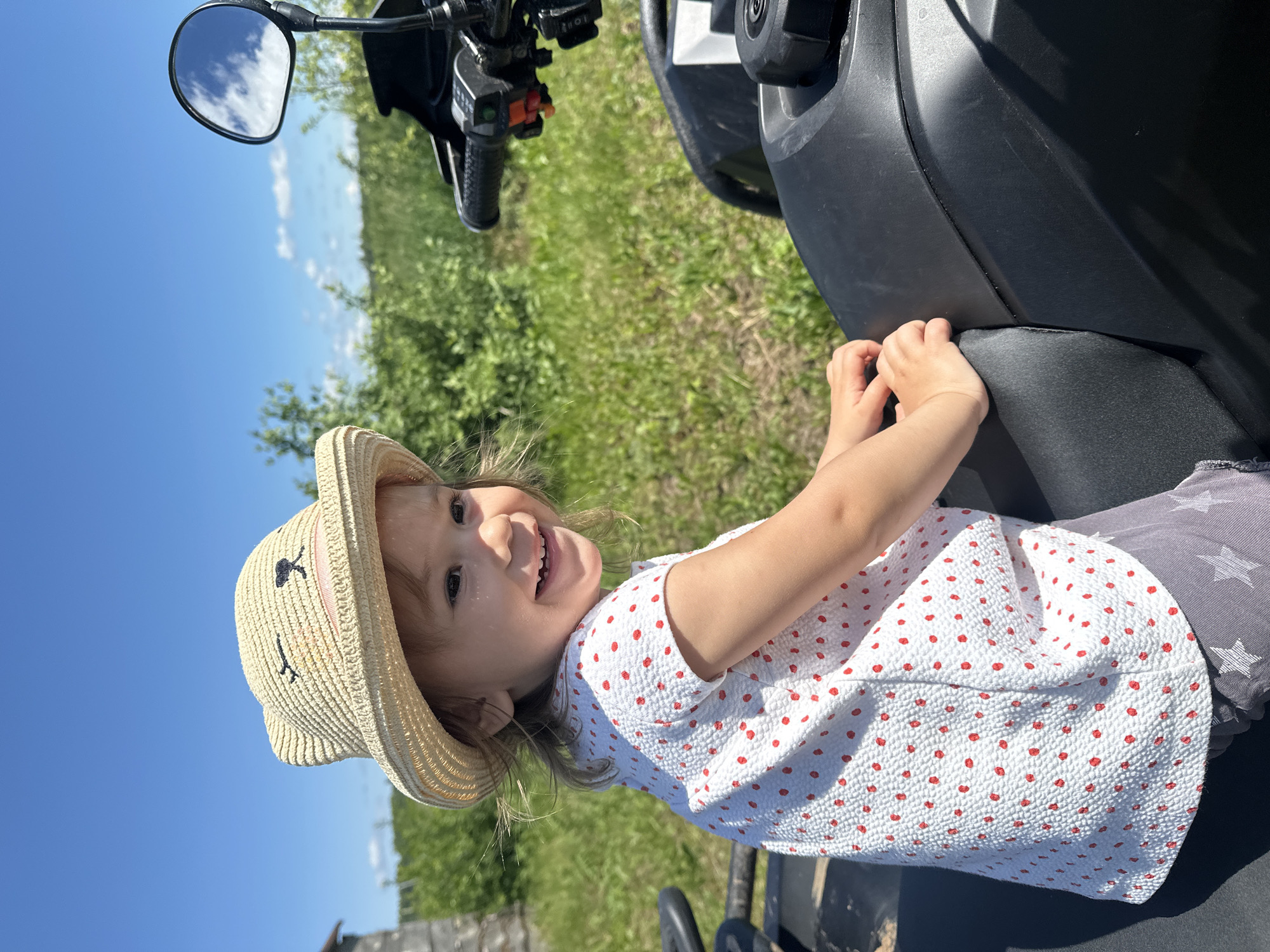 A young Caucasian girl, appearing to be around 2-3 years old, sits on the seat of a black ATV. She is wearing a straw hat with a bear face design, a white textured short-sleeved shirt with red polka dots, and grey pants with white star patterns. Her left hand is resting on the ATV's seat. The girl is smiling broadly and looking to her right. The ATV is parked outdoors in a grassy area with green foliage and trees in the background under a clear blue sky. A rustic wooden structure is visible to the far left. The scene is likely in the village of Kharnavy, Russia, given the natural, rural setting.