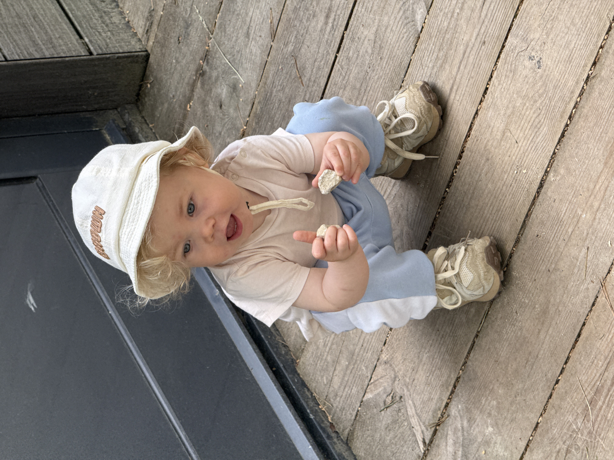 The image features a cheerful baby sitting on wooden decking outdoors, likely on a porch or patio in Dobrograd, Russia. The baby, with fair skin and light hair, looks directly at the camera with wide blue eyes and an open mouth, suggesting a smile, laugh, or curious expression.
The infant is dressed in a light beige short-sleeved t-shirt, light blue pants with a white scalloped trim, and light beige sneakers with white laces. The baby wears a light-colored bucket hat, which has visible text embroidered in a brown/gold, blocky font spelling "DOBROGRAD."
In both hands, the baby holds small, irregularly shaped, light-colored objects, which appear to be pebbles or small pieces of a natural material. The wooden decking beneath the baby is made of worn planks with visible grain, and small bits of debris like pine needles are scattered between the gaps. To the left, a dark, possibly black or dark grey, surface is visible, which could be part of a building structure or door. The scene is well-lit, indicating it is daytime. The overall emotion conveyed is one of joy and curiosity, capturing a moment of a child exploring their surroundings.