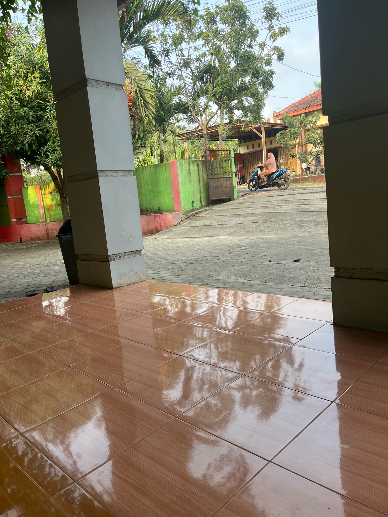 In the city of Kemuning, Indonesia, the image captures a view from under a covered porch with tiled flooring. In the background, a person wearing a pink headscarf and orange top rides a blue motorcycle. The scene includes lush green trees, a partially visible building with a red-tiled roof, and a vibrant green and red painted wall. A black trash bin is positioned near a large support pillar on the left, and a pair of flip-flops rests on the tiled ground in front of it. The overall environment suggests a residential or institutional setting on a clear day.