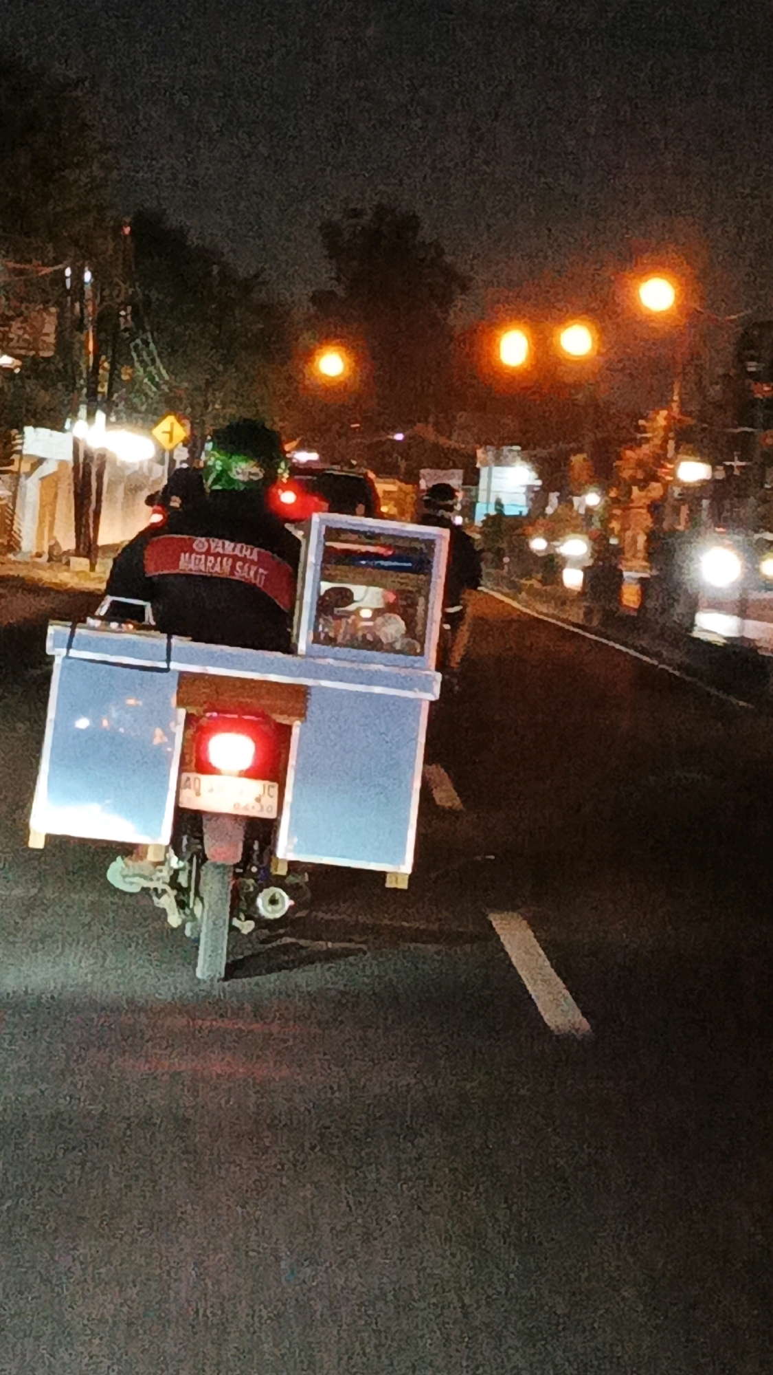 A rear view of a motorcycle is shown at night on a city street in Surakarta, Indonesia. The motorcycle is carrying a large, light blue rectangular box on the back, with a smaller, reflective-sided box mounted on top of the larger one. Inside the smaller box, there appears to be a display of small, round objects. The motorcycle has a red taillight illuminated, and a license plate that reads "AQ 4310 JC". The rider is wearing a black jacket with a red stripe and "YAMAHA MATARAM SAKTI" written on it, and a green head covering. Streetlights and illuminated buildings are visible in the background. The road is asphalt and shows lane markings.