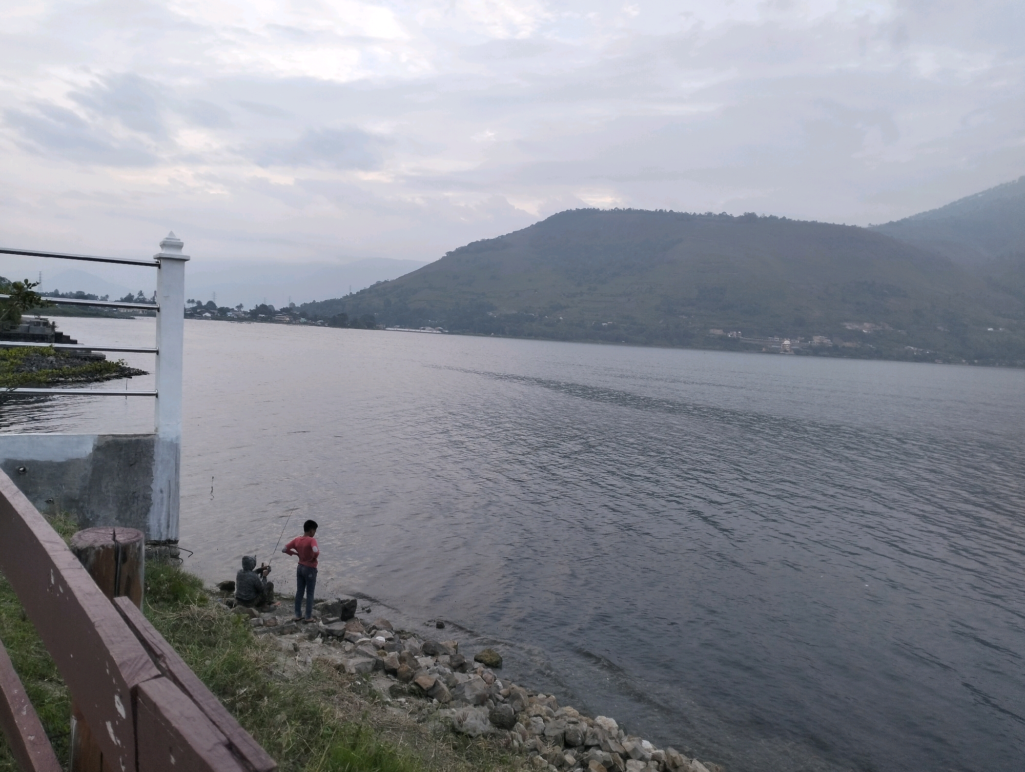 The image depicts a serene lakeside scene with two individuals fishing by the shore. In the foreground, a brown wooden railing partially obstructs the view, with grass and rocks lining the water's edge. A white pillar structure is visible to the left, alongside the water. The lake stretches out, reflecting the overcast sky and the lush, green hills that form the backdrop. The distant hills are shrouded in a light mist, suggesting a cool or humid atmosphere. One person is seated, facing the water, while the other stands, holding a fishing rod. The overall ambiance is tranquil and suggests a peaceful, rural setting in Parlondut, Indonesia, likely during a quiet time of day, perhaps early morning or late afternoon, given the diffused light.