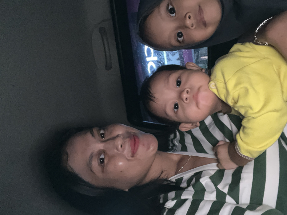 An adult female, an infant, and a young child are captured in a close-up photograph taken inside a vehicle at night in Tambakromo, Indonesia. The adult female, positioned on the left, has dark hair, wears a green and white striped long-sleeved shirt, and a thin silver necklace. She displays a slight smile. She is holding an infant, centrally located, who wears a solid yellow long-sleeved top and a light-colored bracelet on the left wrist, looking directly forward with a neutral expression. To the right of the infant, a young child with dark eyes is present, wearing a dark-colored hijab and a silver-toned bracelet on the left wrist, also looking towards the camera with a neutral expression. The vehicle's interior headliner, light grey in color, is visible at the top, along with a dark grey grab handle. Through the vehicle's rear window, reflections of external artificial lights and a partial, inverted commercial sign reading "OPPO" are discernible, indicating an urban setting.