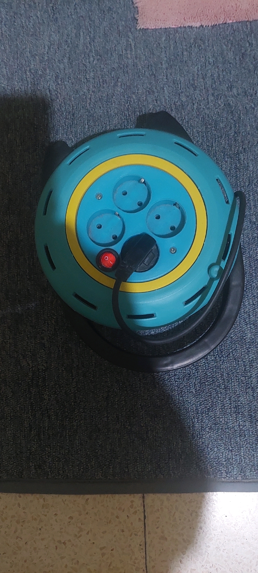 An overhead, medium shot captures a teal and yellow circular extension cord reel resting on a dark blue textured rug. The reel has a prominent yellow band and three circular outlets, each with three pinholes. A black power cord plugs into the center of the reel. A small red on/off switch is positioned to the left of the power cord. The reel has small, horizontal slots along its circumference. The background consists of the rug, with a hint of a pink object in the upper right corner and a transition to a speckled, light-colored floor at the bottom of the frame. There are no people visible in the image. The scene appears to be indoors, possibly a home or office, and the presence of the extension cord suggests an electrical setup. The location of Lumajang, Indonesia, is not visually represented in the image and cannot be inferred.