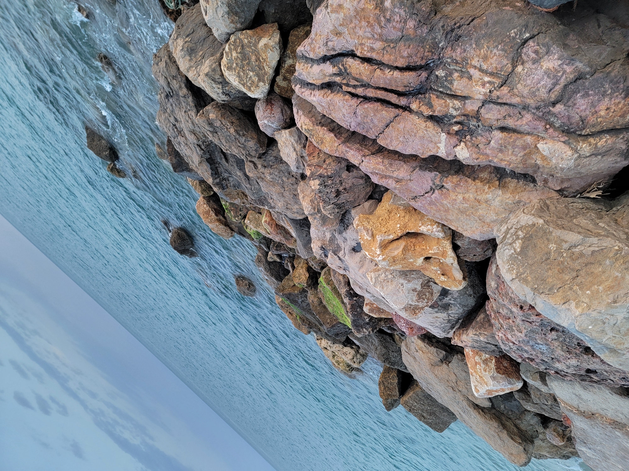 The image depicts a rocky coastline with the ocean meeting the shore. The rocks are of various sizes and colors, ranging from dark gray to reddish-brown and some are covered in patches of green moss. The sea is a vibrant blue-green with gentle waves lapping at the shore, creating a textured surface. The sky above is overcast with light gray clouds stretching across the horizon. The scene is shot at an angle, giving a sense of depth and perspective, with the foreground dominated by the rugged rocks and the background showing the vast expanse of the ocean and sky. This appears to be a natural or semi-natural rocky breakwater or shoreline, potentially in an area like Quarteira, Portugal, known for its coastline. There are no people or animals visible in the image.