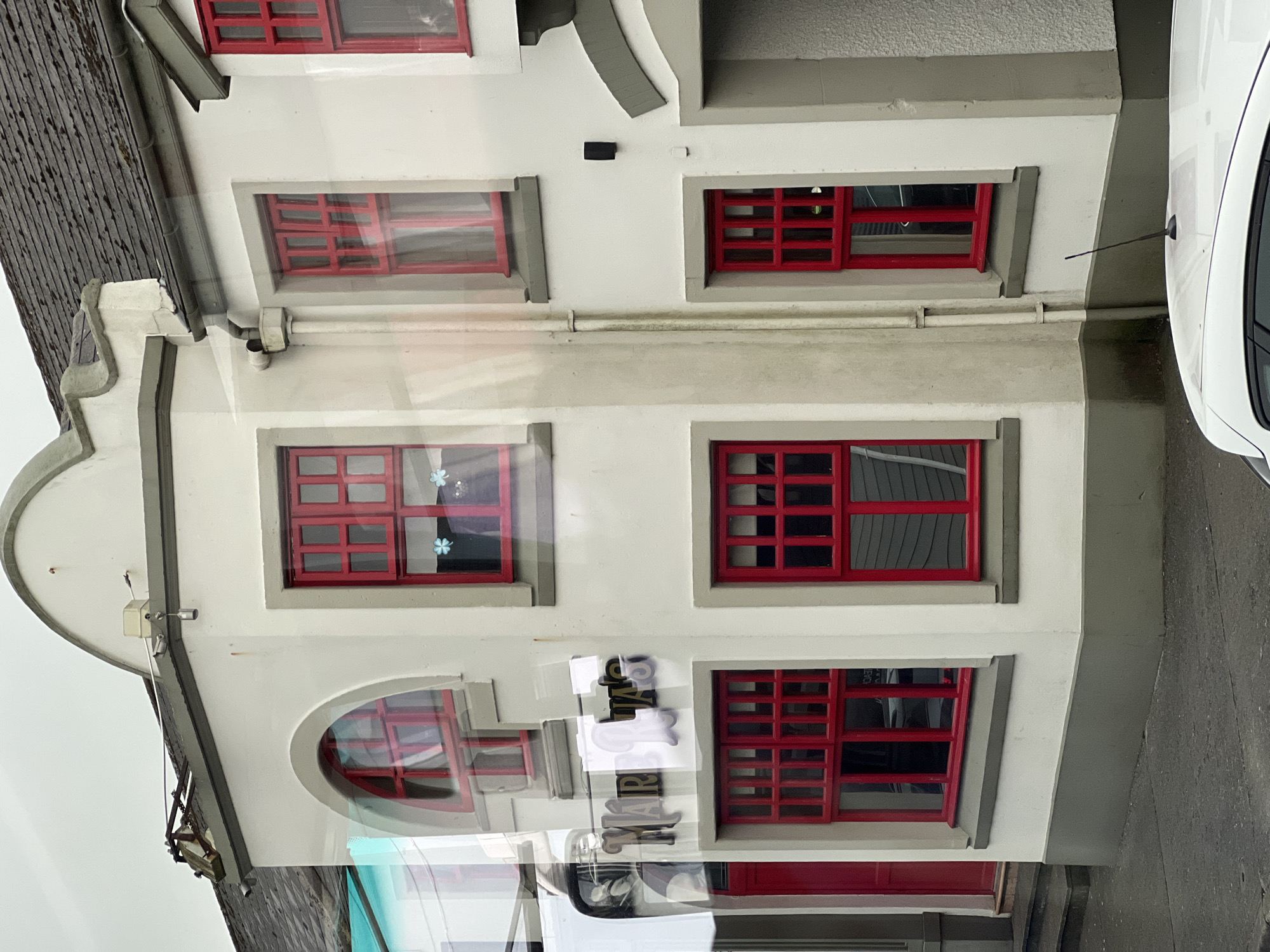An exterior, medium shot captures the facade of a two-story building with white-painted walls and prominent red window frames. The building features several windows of varying sizes and shapes, all with the same red framing. On the ground floor, a red door is visible on the left side, with a small set of concrete steps leading up to it. The building has a dark, tiled roof. A sign with stylized text reading "MARIA'S" is mounted on the facade between two windows. Reflections from what appears to be a bus or vehicle are visible on the building's surface, obscuring some details of the windows and interior. A portion of a white car's roof is visible in the foreground at the bottom of the image. The scene appears to be located in a town or city, with a paved street in front of the building. The sky is overcast and gray.