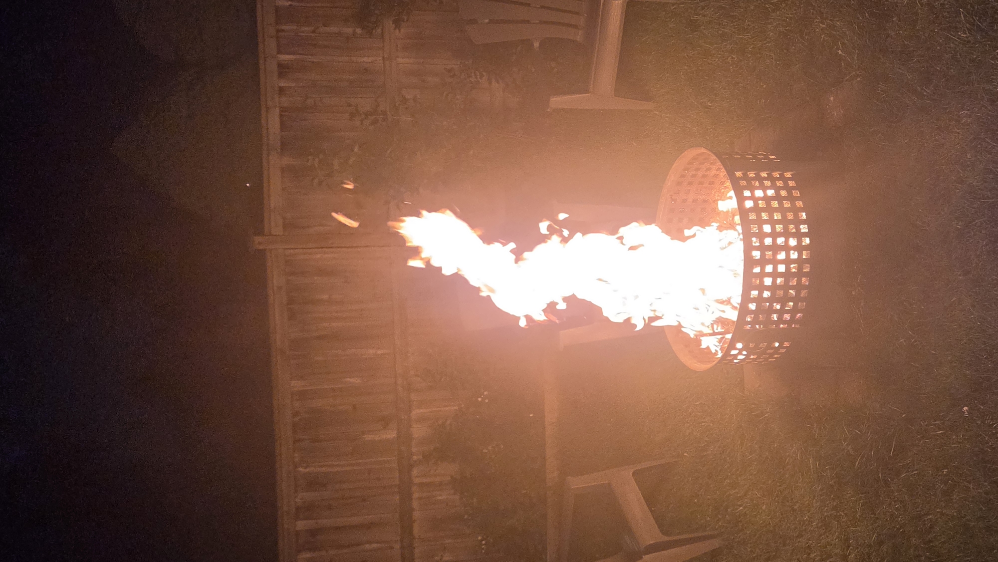 A fire pit is burning brightly in a backyard at night. The fire is tall and orange with some white at its core. It is contained within a circular, metal fire pit with a checkered pattern. The fire pit sits on a patch of grass. To the left and right of the fire pit are two light-colored chairs. Behind the fire pit is a wooden fence. The sky above is dark. There is a tiny white dot in the sky. The scene is illuminated by the fire and appears to be a residential backyard. There are no people visible in the image. The image does not provide any specific information to determine the location within the city of Toronto, Canada.