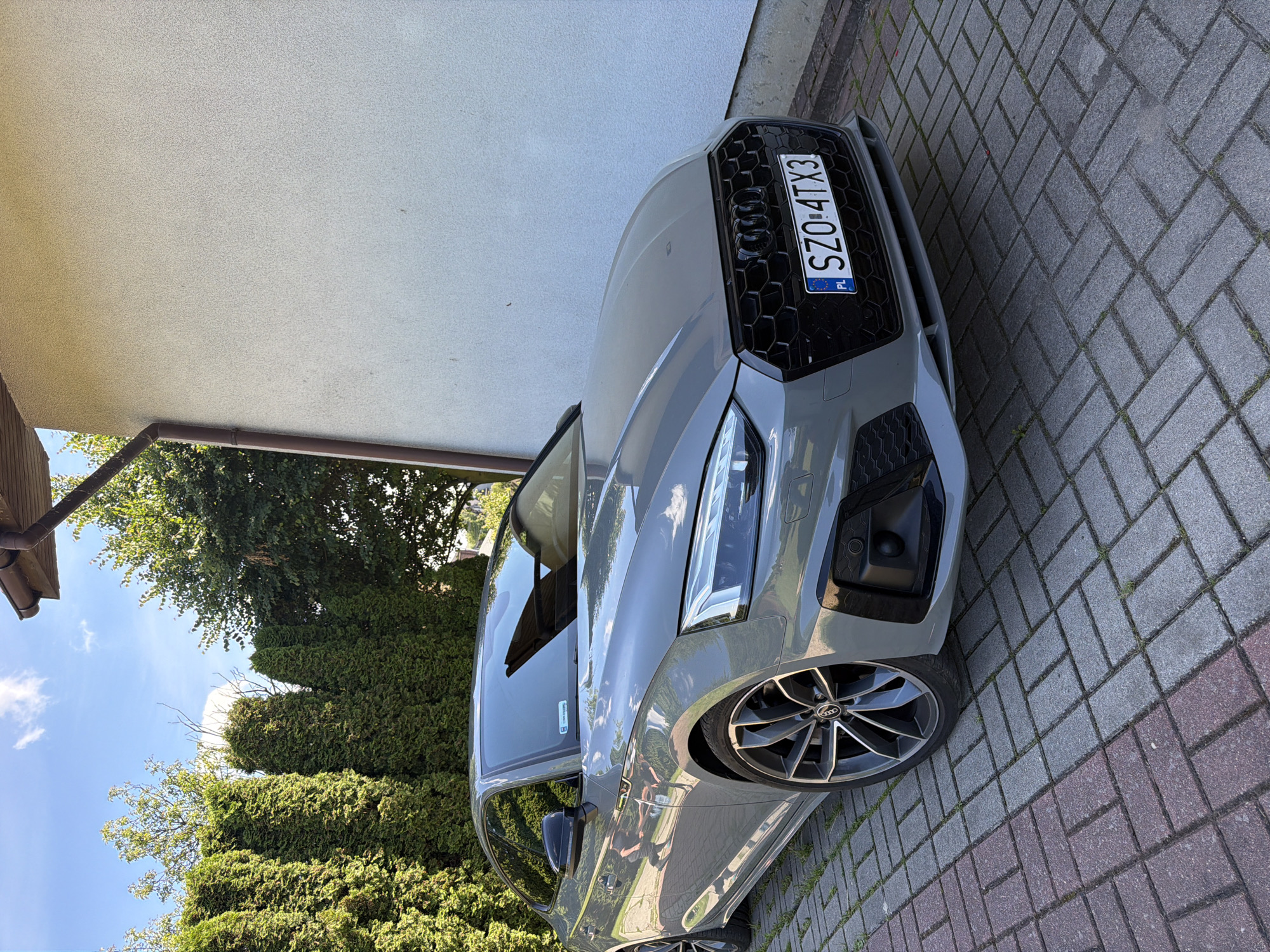 A grey Audi A5 coupe is parked on a herringbone-patterned brick driveway in front of a light-colored stucco house. The car's front is angled towards the viewer, with its license plate reading "SZO 4TX3". A row of tall green evergreen trees lines the left side of the image, and the sky above is blue with scattered white clouds. A gutter downspout is visible on the right side of the house. The scene appears to be a residential exterior setting.