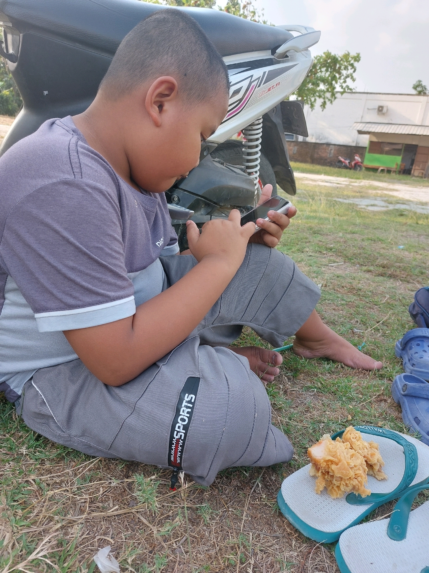A young boy, seated on the ground next to a motorcycle, is engrossed in his smartphone. He is wearing a grey t-shirt with lighter grey accents and grey shorts with the word "SPORTS" printed on a black tag. His bare feet are visible as he sits on the dry grass. In front of him, on a pair of light blue flip-flops, is a serving of fried chicken. The background shows a building with an air conditioning unit and a fence, indicating an outdoor, possibly rural or semi-urban, setting in Sarangan, Indonesia. The lighting suggests it is daytime.