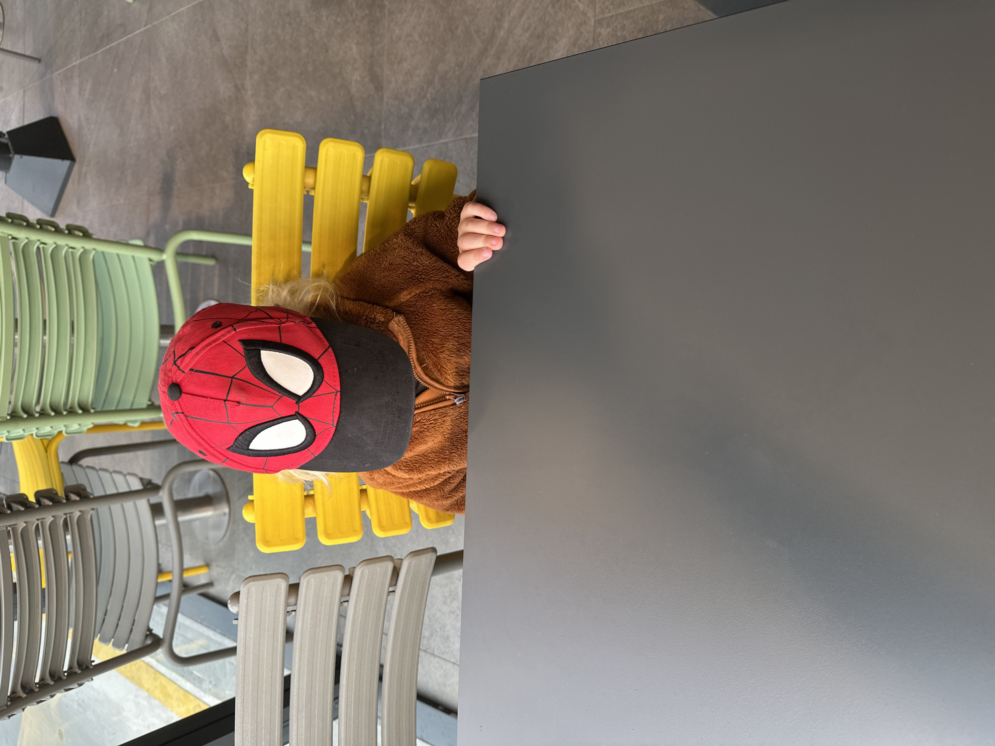 A top-down shot reveals a child wearing a red and black Spider-Man hat and a brown fleece jacket sitting at a yellow slatted chair. The child's right hand is visible on the edge of a grey table that takes up a significant portion of the right side of the frame. A green slatted chair is partially visible to the left of the yellow chair, and a grey slatted chair is partially visible below. The floor appears to be grey tile. The location is Fethiye, Turkey.