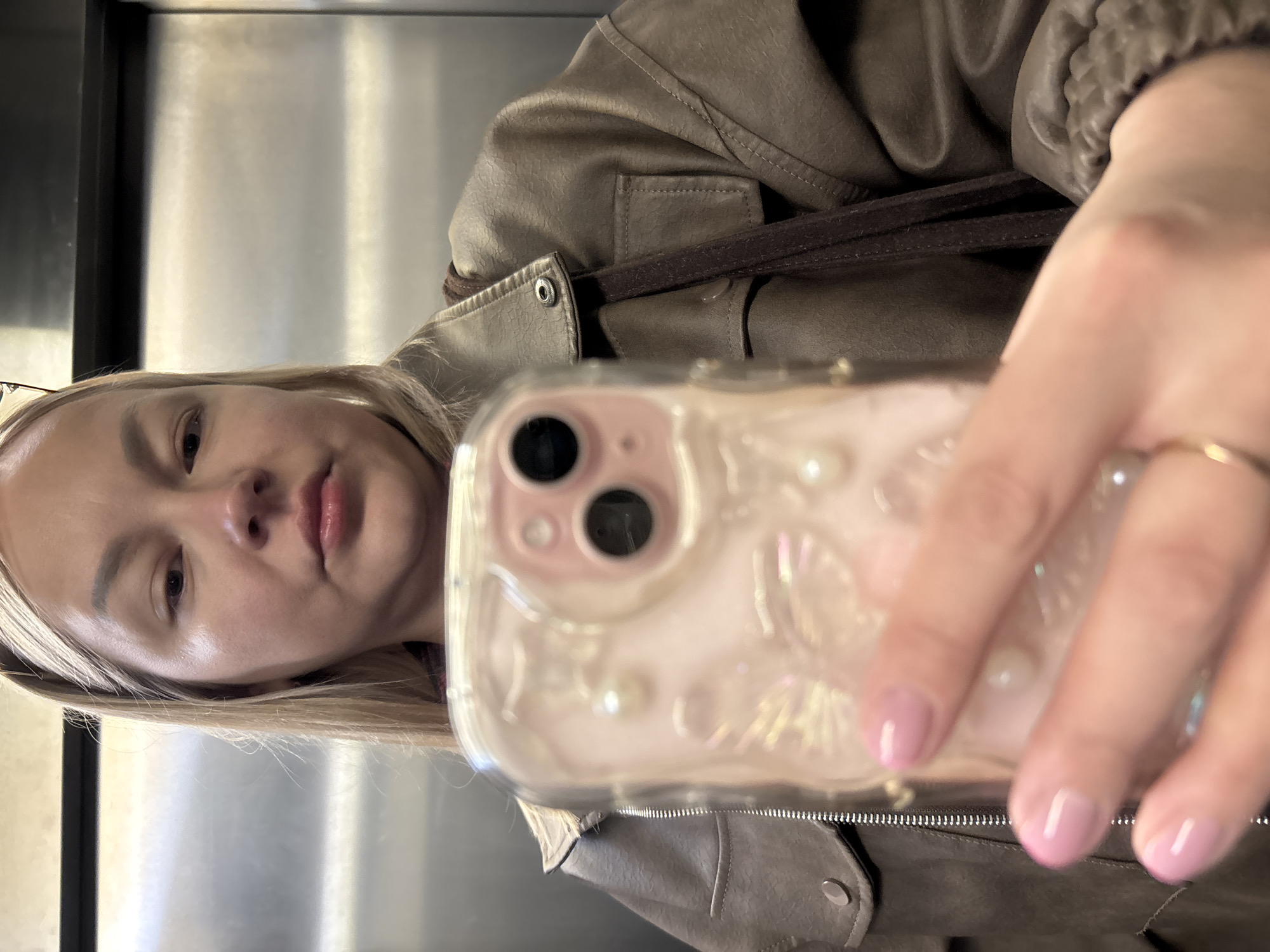 A woman is taking a selfie in what appears to be an elevator. She has blonde hair and is wearing a brown leather jacket with a brown strap. She is holding a smartphone in front of her face. The phone has a clear case with pearlescent detailing. Her nails are painted a light pink. The elevator appears to have stainless steel walls. The location is unknown and cannot be determined to be Izhevsk, Russia from the image.