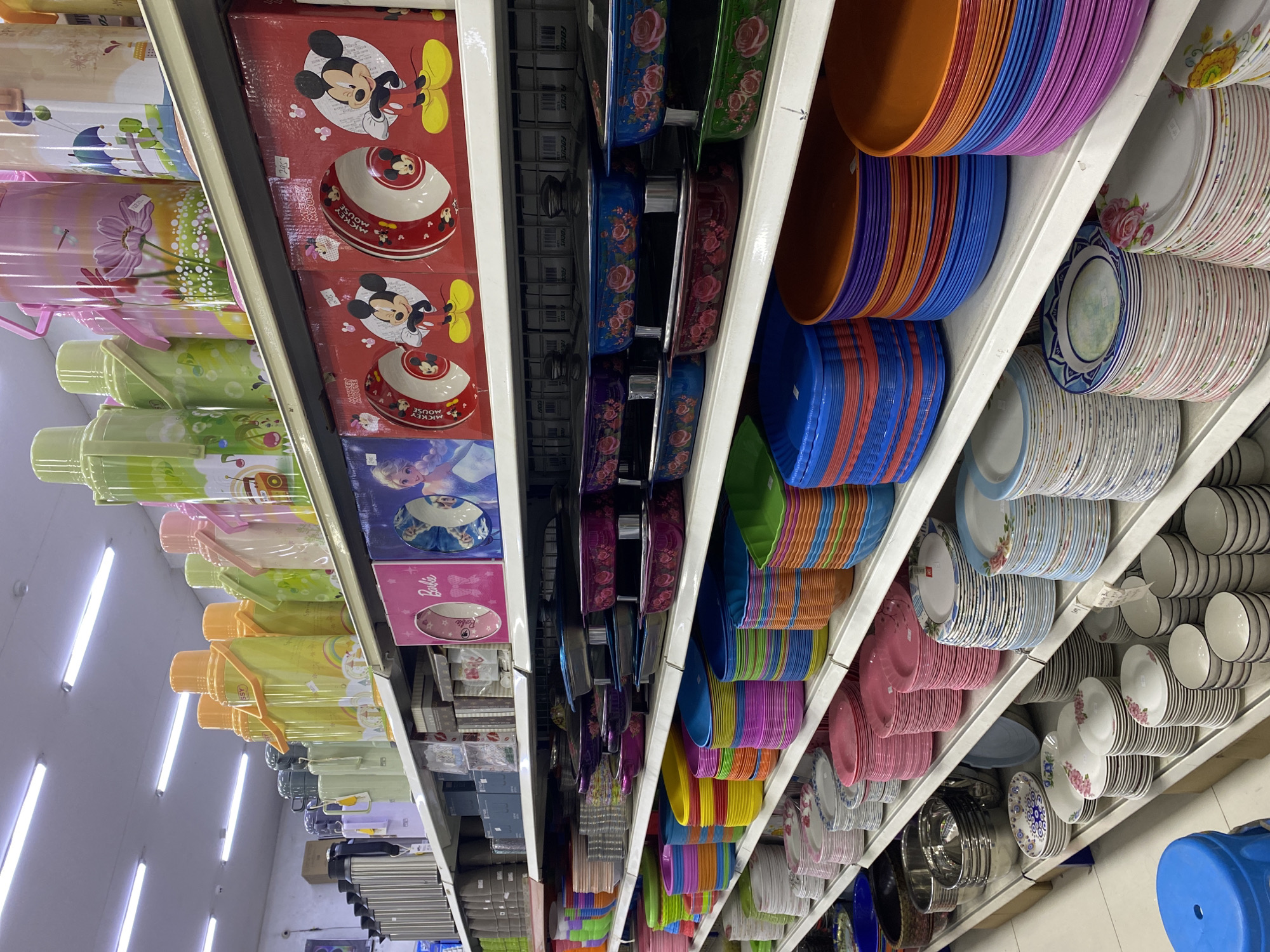 The image depicts a retail store shelf display, likely in Davao City, Philippines, filled with an assortment of kitchenware and household items. The shelves are organized with various products, including stacks of colorful plastic plates, bowls, and platters in a wide spectrum of hues such as blue, orange, purple, pink, and green. There are also metal thermos containers, decorative boxes, and packaged dinnerware featuring cartoon characters like Mickey Mouse and Disney princesses. The lighting suggests an indoor environment, possibly a department store or a specialty shop. The scene is static, with no people visible in the frame.