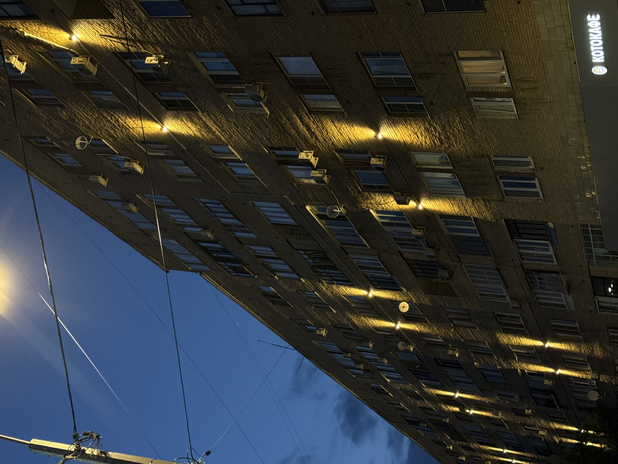 The image shows a multi-story brick apartment building at night, illuminated by upward-facing lights installed along its facade. The sky is a deep blue with some clouds. Power lines and a street lamp are visible in the foreground, with one light prominently overexposing the upper left portion of the image. The building has numerous windows, many with air conditioning units installed outside them. A sign that reads "KOTOKAFE" with a logo is visible at the bottom right of the building. The scene is set in the city of Lobnya, Russia. There are no people visible in the image.