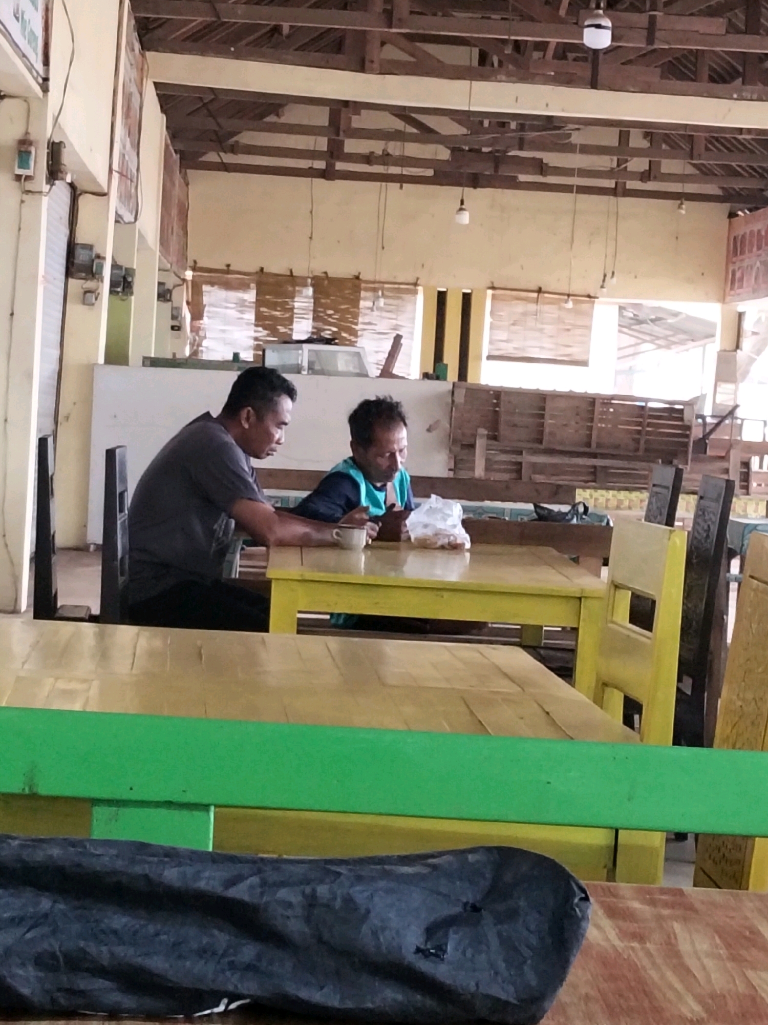Two men are seated at a yellow table in an open-air establishment in Tasikmadu, Indonesia. The man on the left, wearing a dark gray t-shirt and dark pants, is looking down at a white bowl in front of him, holding a spoon. The man on the right, wearing a teal vest over a dark shirt, is looking at a plastic bag of food and a phone. The establishment has wooden beams in the ceiling and light-colored walls. Yellow tables and chairs are present throughout the space. In the foreground, a green railing separates the seating area from another section with wooden tables. A dark, crumpled object, possibly a bag, rests on a table in the lower portion of the image.