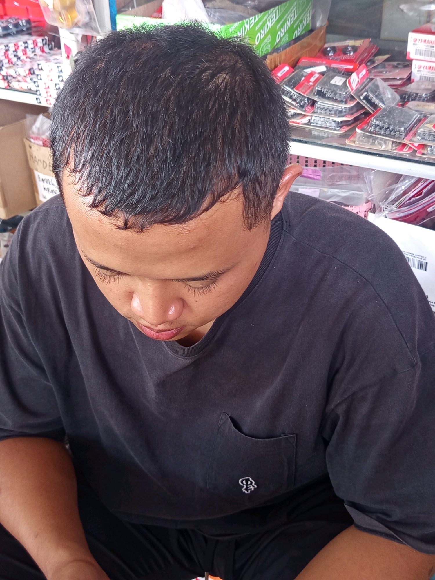 In Karang Sari, Indonesia, a person is seated, looking down at their hands. They are wearing a dark gray t-shirt with a small embroidered logo on the left chest pocket. The background suggests a retail environment, with shelves stocked with various items, including boxes and packaged goods, some of which appear to be motorcycle parts.