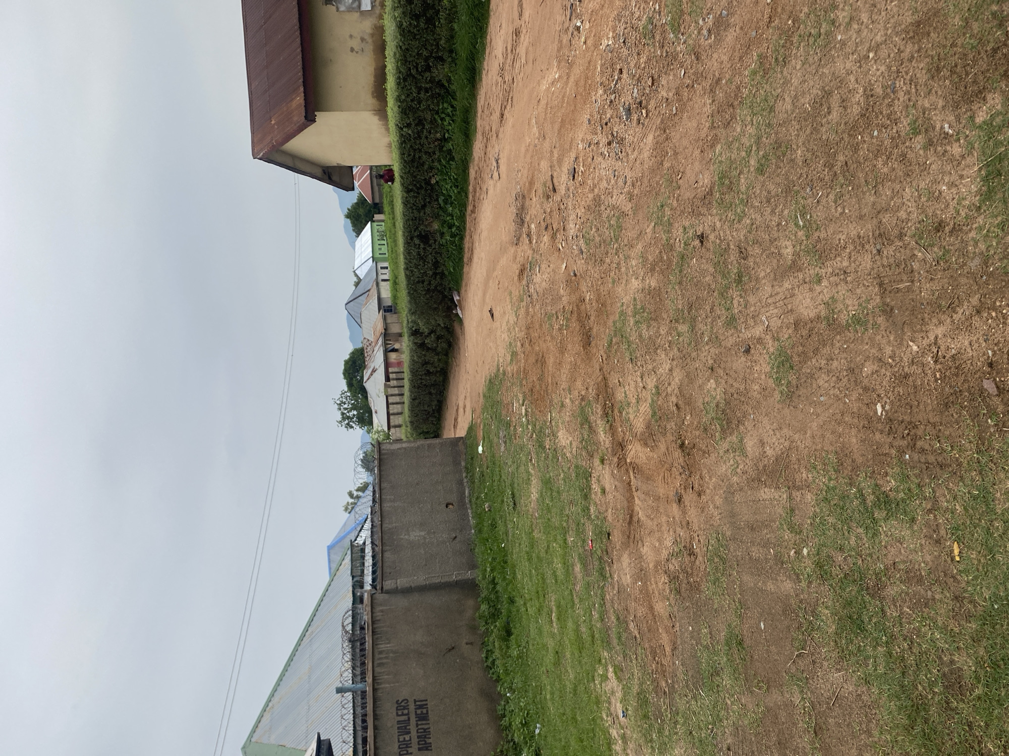 This is a photograph of a dirt road and grassy area with buildings in the background. The scene appears to be outdoors during the daytime, under an overcast sky. The road is unpaved and shows tire tracks, indicating vehicular use. To the left of the road, there is a well-maintained hedge that runs alongside several low-rise buildings with corrugated metal roofs. These buildings have a somewhat rustic appearance, with some showing signs of wear and tear.
Further in the background, more buildings are visible, including one with a lighter colored exterior and a red metal roof. There are also trees and other vegetation scattered throughout the scene. On the left side of the image, a concrete wall is present with visible text that reads "PREVAILERS APARTMENT". Barbed wire is installed along the top of this wall, suggesting a security measure.
The overall impression is that of a semi-developed or rural area. The lighting is flat due to the overcast sky, and there are no clear indicators of a specific time of day beyond it being daylight. The presence of the buildings and the road suggests a residential or commercial area. No people are clearly visible in the photograph, although there might be a small figure in the distance behind the hedge. The weather appears to be cloudy and possibly humid, common in tropical or subtropical climates.