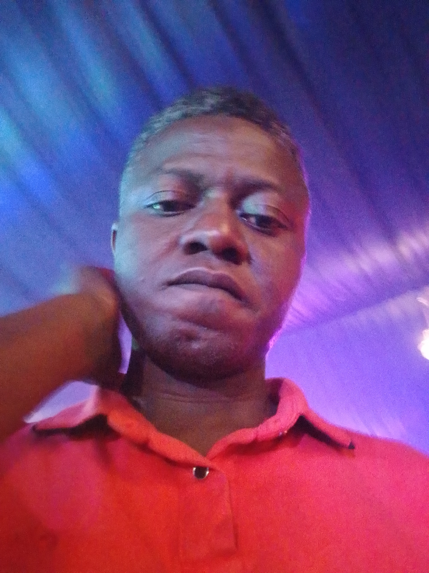 The image is a close-up portrait, likely a selfie, of an adult male. He has dark skin and short, closely cropped hair. He is looking directly into the camera with a neutral to slightly serious expression. He wears a red polo shirt with a two-button placket; a single silver-colored button is visible at the top. His right arm is visible on the left side of the frame, with his hand positioned near his jawline or neck.
The background features strong artificial lighting in vibrant shades of blue and purple, with discernible linear patterns that suggest spotlights or decorative lights. This ambient lighting casts a subtle purplish hue onto parts of the man's face and shirt. The setting appears to be an indoor environment, possibly an entertainment venue like a club or bar, indicated by the atmospheric and colored lighting. The image exhibits some digital noise, common in low-light photography. There is no visible text in the image. The provided location context for the image is Rido, Nigeria.