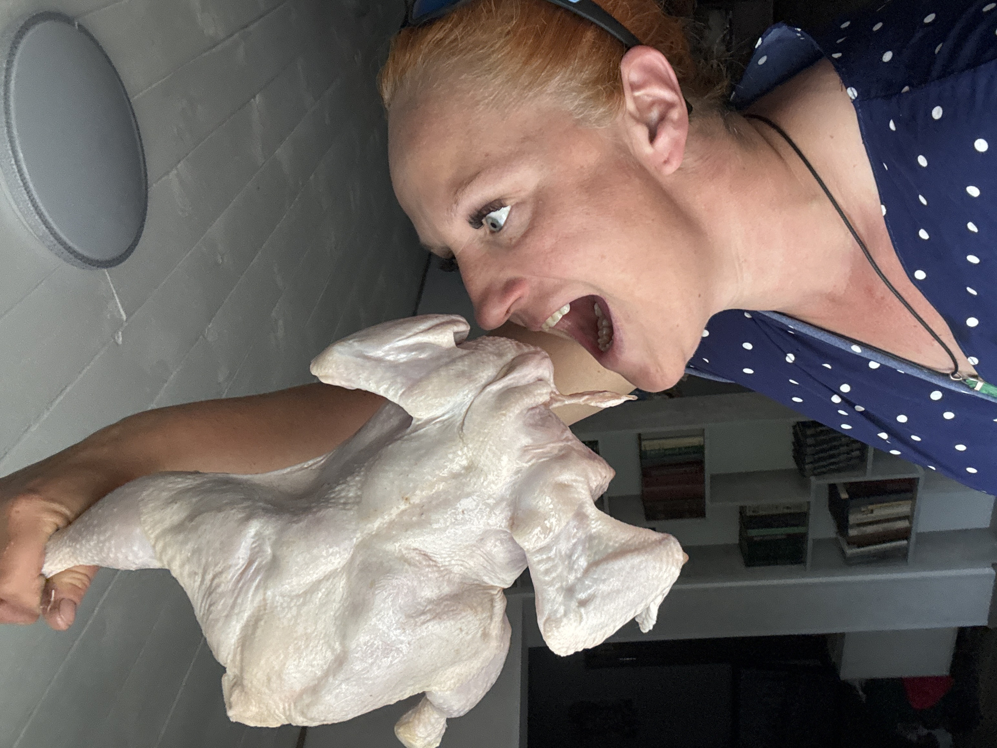 A woman with red hair is photographed in extreme close-up, her face contorted into an expression of mock terror or exaggerated disgust. She is wearing sunglasses on her head and a blue, white polka-dot top. She is holding a raw chicken in her left hand, bringing it close to her open mouth as if she is about to bite it. The background includes a gray wall with a circular light fixture and shelves with stacked items. The location is a building in the city of Chaikovsky, Russia.