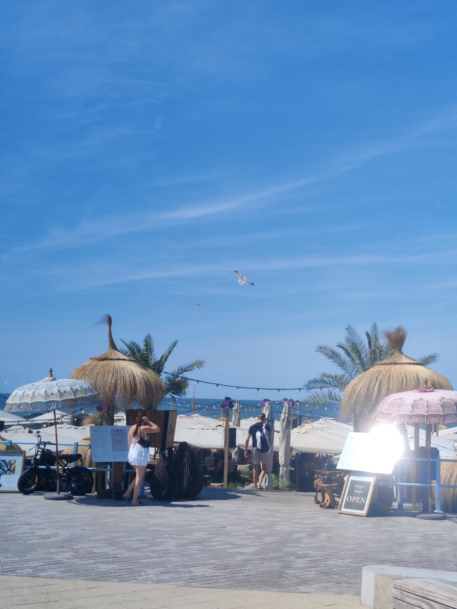This is a photograph of a beachside establishment in The Hague, Netherlands, under a clear blue sky with scattered clouds. The scene depicts a paved walkway leading to a beach area furnished with thatched umbrellas, palm trees, and beach chairs. In the foreground, a black electric scooter is parked next to a stand displaying a menu. A woman in a white dress is standing by the menu, and a man wearing a t-shirt and shorts is walking away from the camera towards the beach. A sign on an easel reads "OPEN." The background shows the sea and more beach umbrellas. Seagulls are visible flying in the sky.
