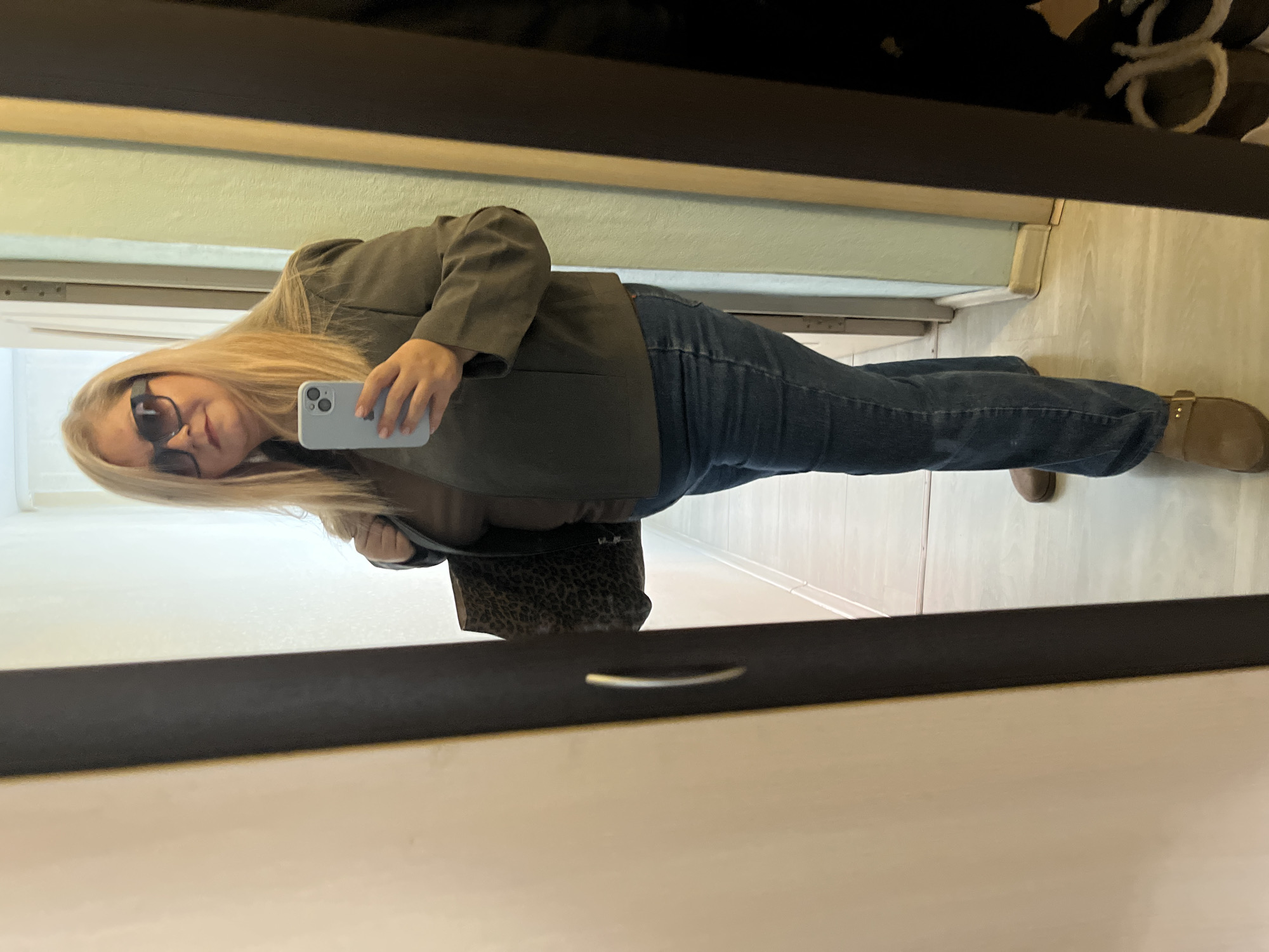 A person is standing in front of a mirror, taking a picture with their phone. The person is wearing sunglasses, a green blazer, blue jeans, and brown sandals. They are also holding a leopard print purse. The background appears to be a hallway with a door on the left. There is no indication of Izhevsk, Russia.