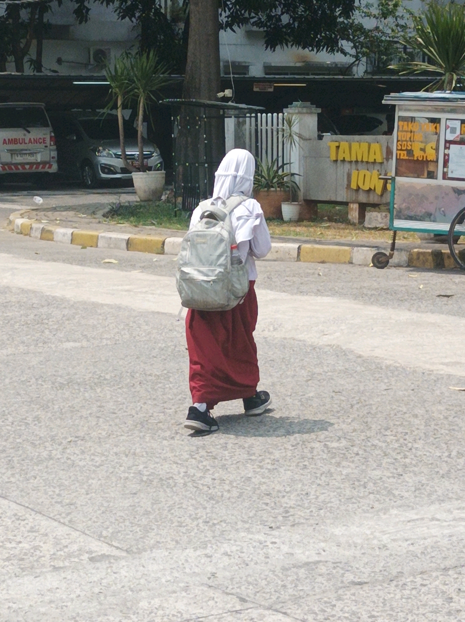 A person wearing a white hijab, white top, and a maroon skirt walks away from the camera on a paved surface in West Jakarta, Indonesia. The person is carrying a light gray backpack. In the background, there is an ambulance with "AMBULANCE" written on its side, a silver car, a sign with yellow lettering that reads "TAMA 104", and a street vendor's cart. Trees and buildings are also visible.