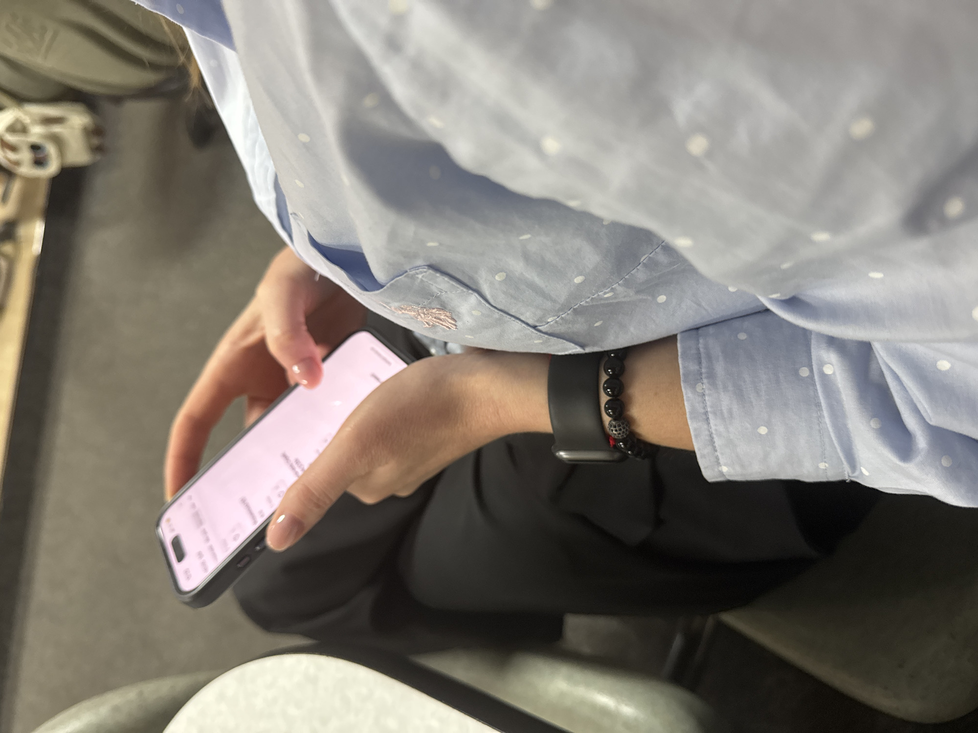 A person wearing a light blue, polka-dotted button-down shirt and black pants is holding a smartphone. An Apple Watch and a beaded bracelet adorn their left wrist. The smartphone's screen is on, displaying text. The setting appears to be indoors, possibly in a public transport vehicle, as suggested by the metallic and upholstered surfaces visible in the background. The image provides no evidence to confirm or deny the location in the city of Kharkiv, Ukraine.
