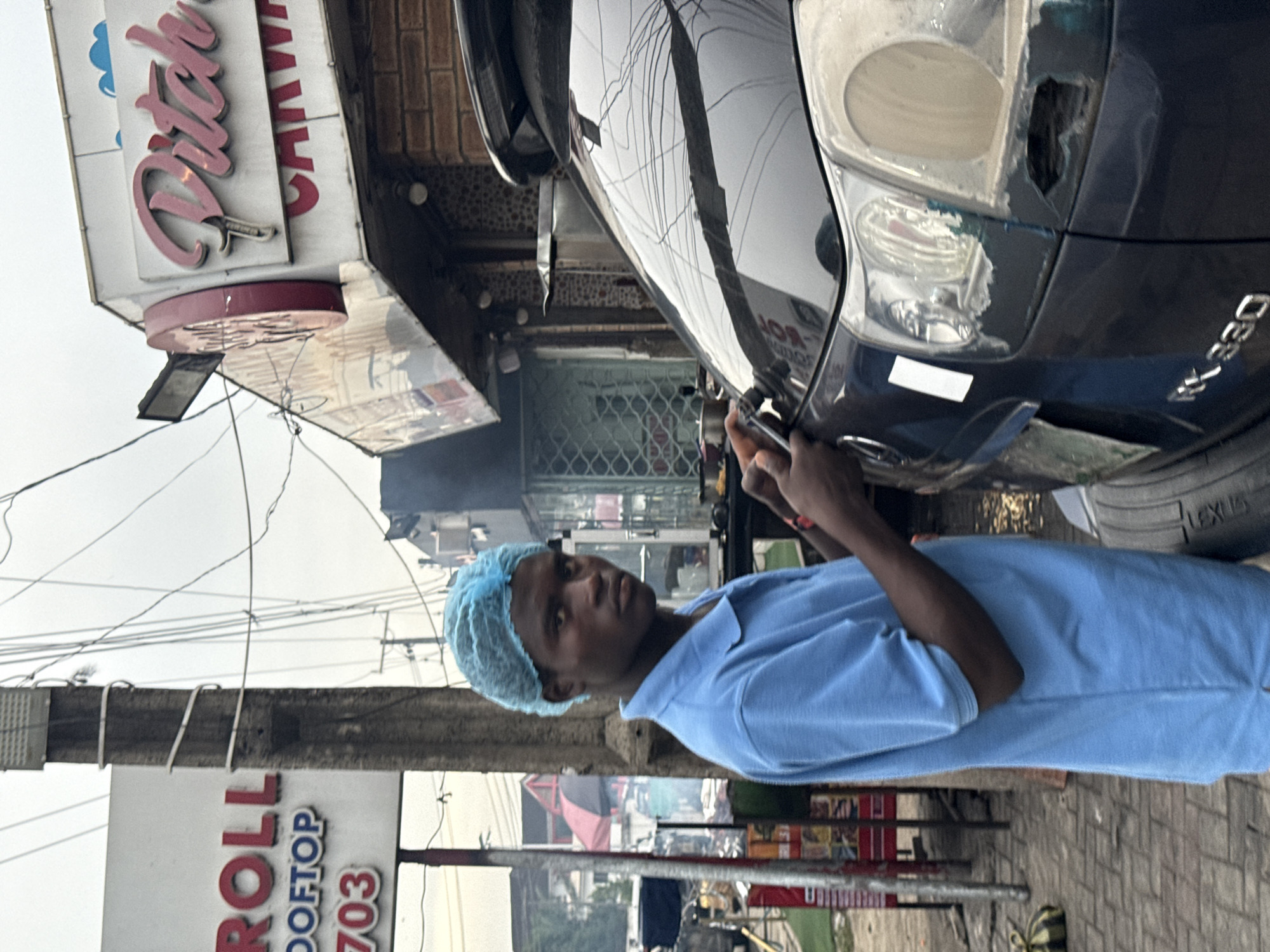 A medium shot depicts a man in a light blue uniform and hairnet, apparently working on a black Lexus RX330 car with a damaged tail light. The scene is set in Ado, Nigeria, suggested by signage in the background displaying "ROLL ROOFTOP 703" and "Ditch Carwash." The man appears to be focused on repairing the car's rear light. The urban environment includes electrical wires overhead, shops, and pedestrian activity.