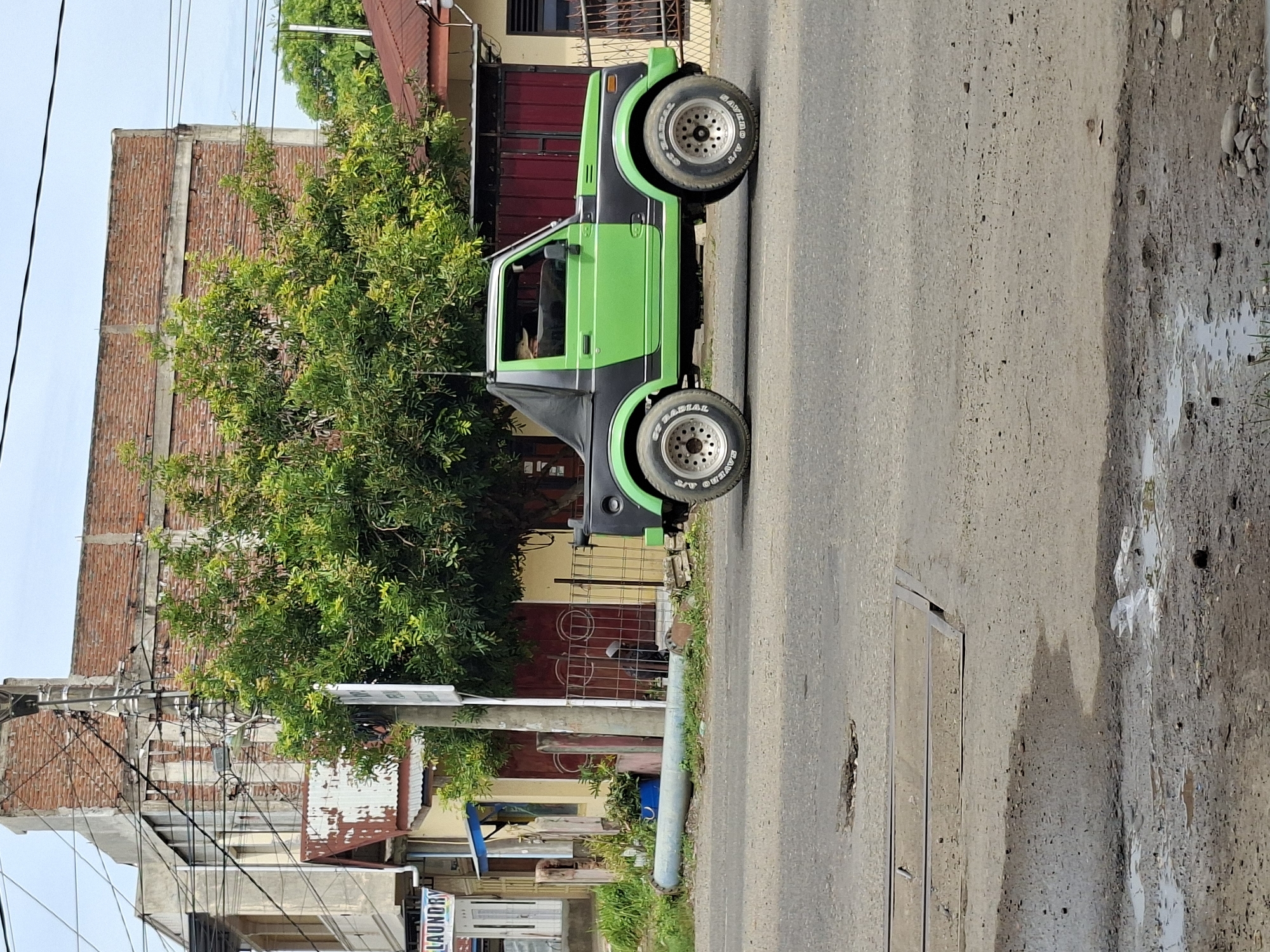 A two-tone green and black Suzuki Samurai is parked on the side of a road in Lhokseumawe, Indonesia. The vehicle has a black soft top and custom wheels. In the background, a brick building with exposed concrete structures and a weathered metal roof is visible, along with dense green foliage. The street is paved and shows signs of wear, including a pothole and puddles of water. Utility poles and wires are present above the scene, indicating an urban or semi-urban environment. There are no people clearly visible in the immediate vicinity of the car, though a shadowy figure can be seen through the driver's side window.