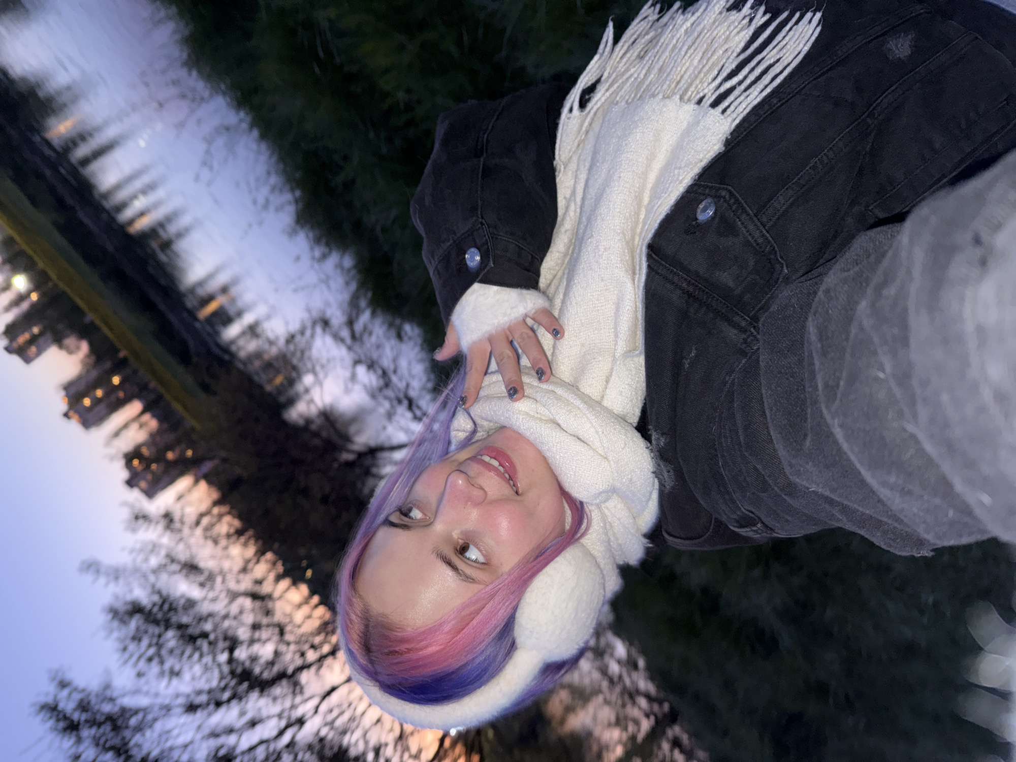 A woman wearing earmuffs and a scarf is the focus of the photograph. The background features a body of water reflecting city lights and bare trees. The location is in the City of Zagreb, Croatia.