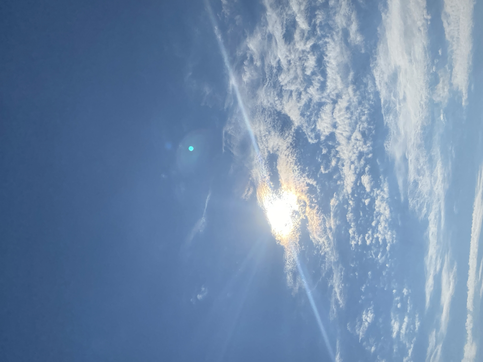 The image shows a daytime sky in Malyi Bukor, Russia. The sky is primarily blue with a mix of scattered cumulus and cirrus clouds. The sun is visible, partially obscured by the clouds, creating a bright glare and sunbeams. The image is angled, with the clear blue sky on the left and the clouds concentrated on the right. There are no people or objects visible in the foreground or midground. The environment consists solely of the sky and clouds.