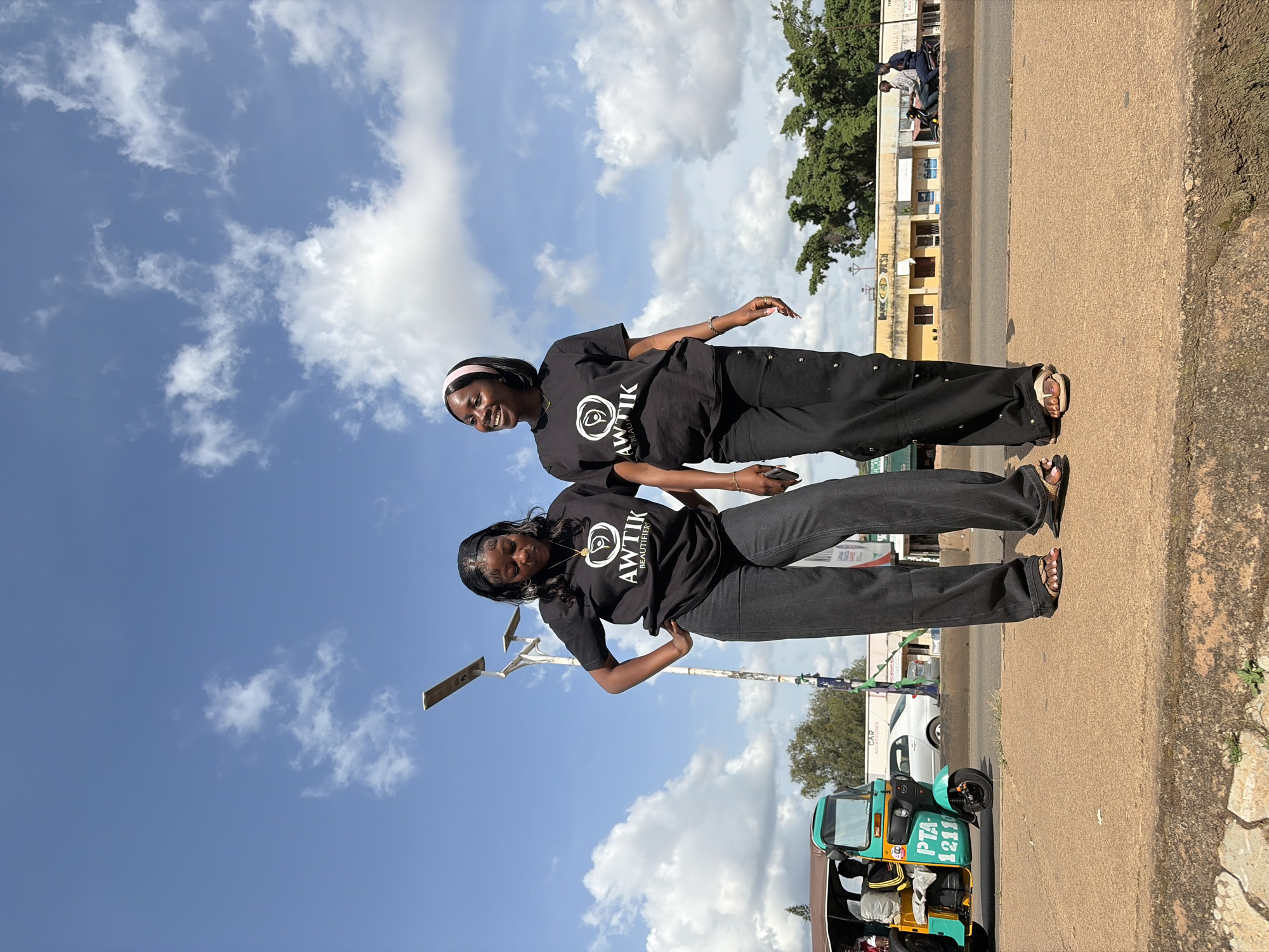 Two young Black women stand on a raised concrete sidewalk in Gura Top, Nigeria, under a bright blue sky dotted with white clouds. Both are wearing black t-shirts with a white logo that reads "AWTİK BEAUTIFIERS" and black, wide-leg pants. The woman on the left has her hands on her hips and is looking down. The woman on the right is looking up and to her left, smiling. She has a pink headband in her hair.
Behind them, a road is visible with a turquoise and yellow auto-rickshaw, also known as a "Keke Napep," with the license plate "PTA 1211" on the front. A white sedan is parked further down the road, and several buildings with yellow and white facades line the street. Trees with dense green foliage are visible behind the buildings. A lamppost with two lights stands between the two women. The overall scene depicts a typical street view in a Nigerian city.