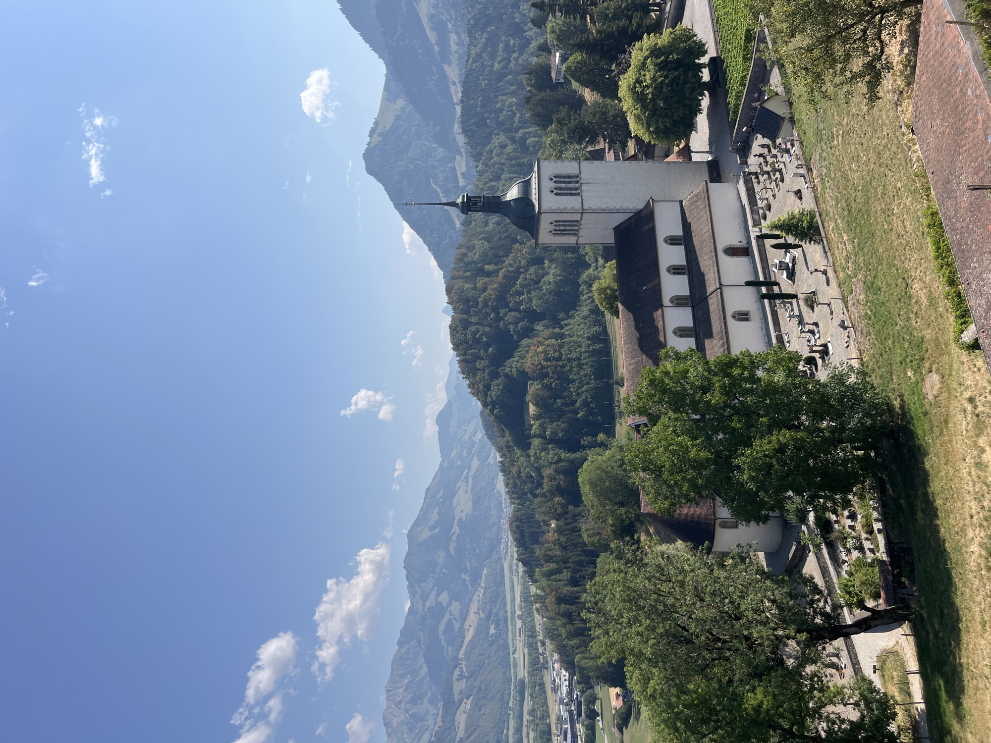 The image shows a wide-angle view of a church with a tall, slender steeple, set against a backdrop of rolling, tree-covered hills and distant, hazy mountains under a clear blue sky with scattered white clouds. The church is white with arched windows and a dark, pointed roof on the steeple. Adjacent to the church is a cemetery with numerous gravestones and small crosses, some organized in neat rows, others more scattered. Several mature trees with lush green foliage are present in the foreground and around the church and cemetery.
In the middle ground, the landscape transitions to a valley with more trees, fields, and some buildings suggesting a small town or village. Further in the distance, the mountains are layered and appear to recede into the haze, with some areas showing green fields and possibly more scattered settlements. The overall scene conveys a sense of rural tranquility and historical presence, possibly in a mountainous region.
The location is identified as Epagny, Switzerland. The time of day appears to be midday or early afternoon, given the bright sunlight and clear shadows. There are no people visible in the image, and no specific actions or interactions are depicted. The environment is natural and somewhat pastoral, with a man-made structure (the church and cemetery) integrated into the landscape.