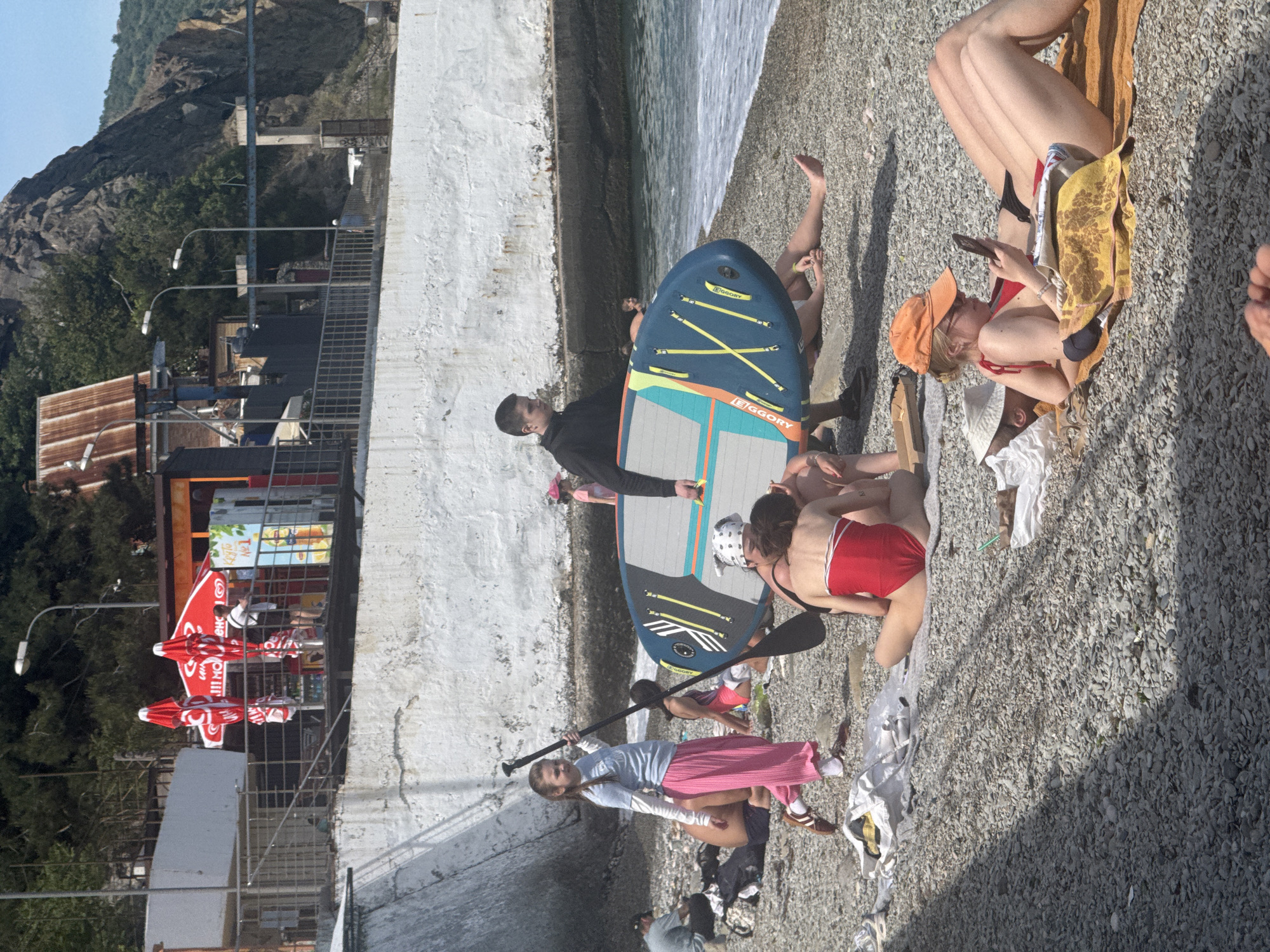 This media file captures a sunny, leisurely scene on a pebble beach, oriented with the sea to the right and a concrete wall/promenade to the left. Several individuals are present, engaged in recreational activities.
In the foreground, a man in a dark top holds a blue paddleboard with prominent yellow straps, featuring the brand name "EGGORY." A young girl in a light blue top and pink pants stands nearby, holding a black paddle. Closer to the water, a person in a red swimsuit is partially visible. Further back, another person in a red bikini and an orange cap lies on a striped towel, engrossed in a mobile phone. Other people are scattered across the pebbly shore, some relaxing on towels.
Along the concrete wall in the mid-ground, there's a beachside establishment, likely a cafe or vendor, characterized by vibrant red umbrellas with visible Cyrillic text, partially reading "!!!МОРОЖЕН" (suggesting "ice cream"). Tables and chairs are arranged under these umbrellas. Beyond these structures, a mountainous or hilly landscape rises, featuring sparse vegetation, some buildings with corrugated roofs, streetlights, and a metal staircase or pathway ascending the incline.
The time of day appears to be bright daylight, indicated by strong illumination and clear shadows. The overall mood is relaxed and recreational, typical of a summer day at the beach.