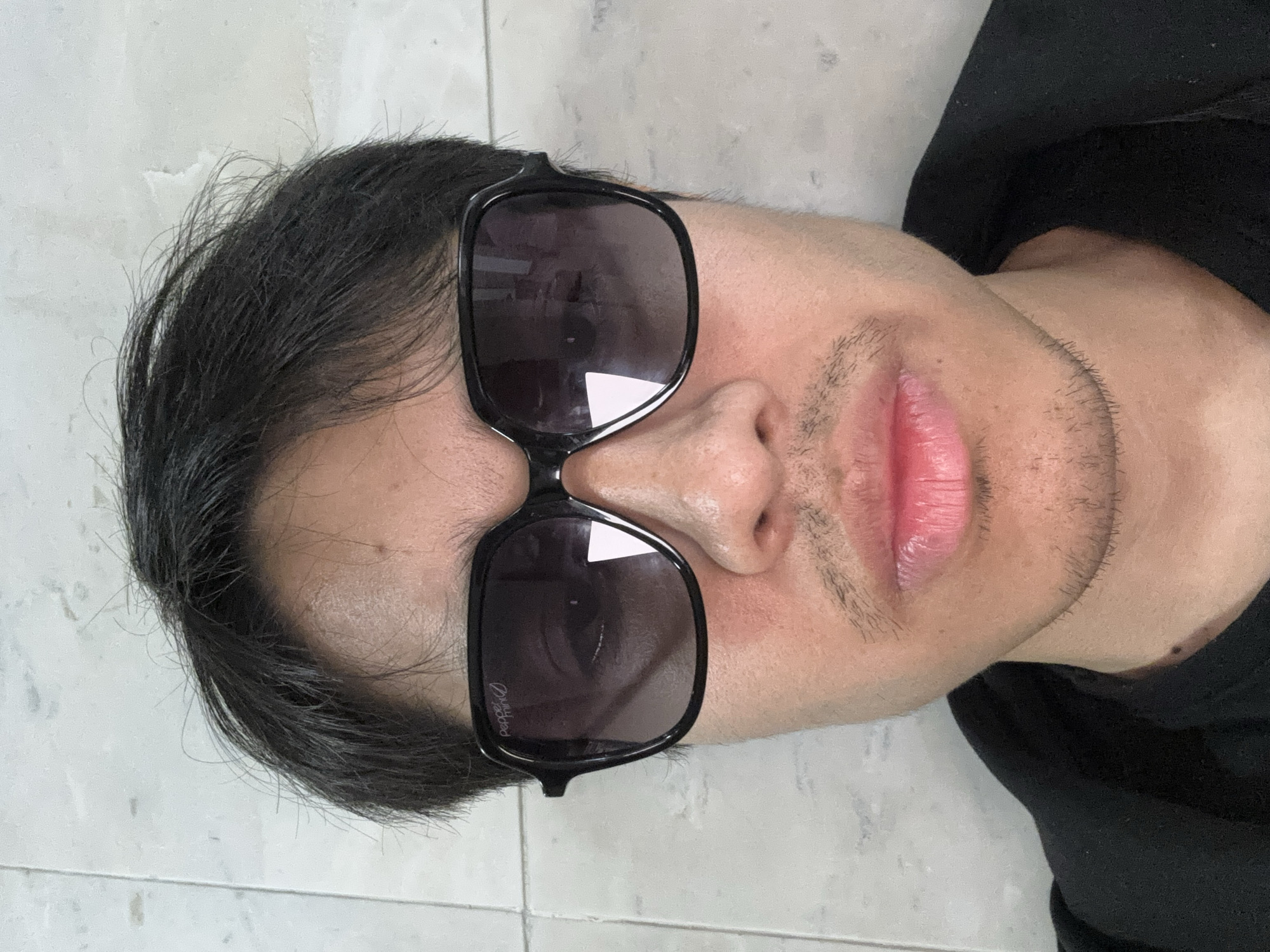 A man wearing sunglasses is lying on a tiled surface. The tiled surface is white. The man has dark hair, a mustache, and some stubble. He is wearing a black shirt. The sunglasses are black with dark lenses. The location is unknown and cannot be determined from the image.