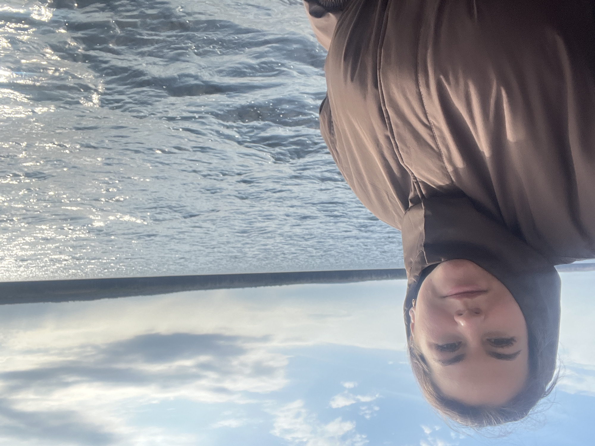 A woman in a brown hooded jacket is shown in a rotated selfie-style photograph. The background includes a body of water with sunlight reflecting off the surface. Beyond the water is a line of trees. Above the trees, there is a partially cloudy sky. There are no specific details suggesting the exact location within the city of Chaikovsky, Russia.