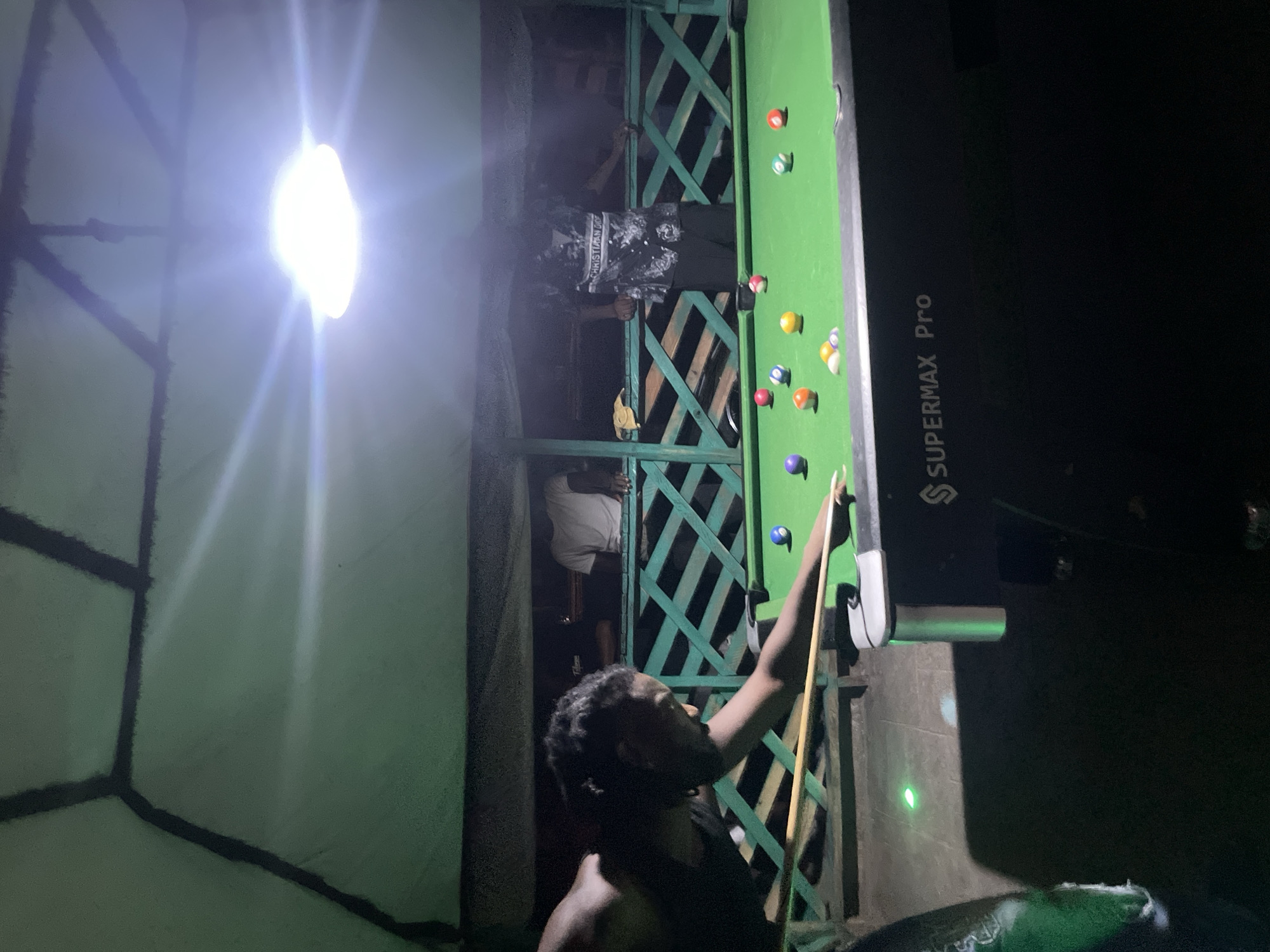 The image depicts a nighttime scene in Lokoja, Nigeria, featuring a billiards game in progress. A man is holding a cue stick, poised to strike a ball on a green-felted "SUPERMAX Pro" pool table. Several billiard balls are scattered across the table. A lattice-like structure forms a backdrop, and other individuals are visible behind it. A bright light source illuminates the scene, creating a strong glare and casting shadows.