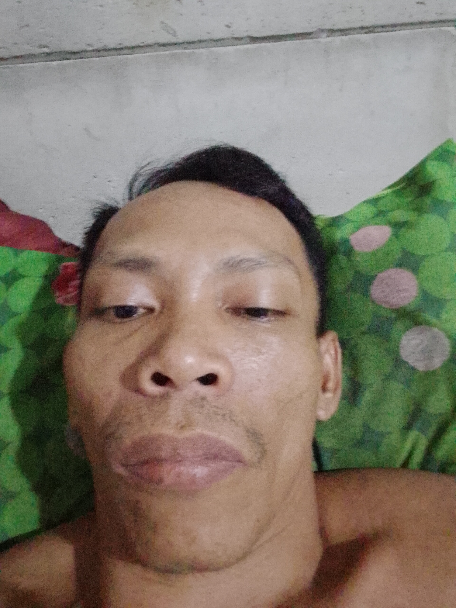 A close-up selfie of a man lying on a green and pink polka-dot pillow or similar surface. The background includes a grey wall or ceiling. The man appears to be indoors, possibly in a residential setting. No specific information suggests the exact location in Bukit Ketok, Indonesia.